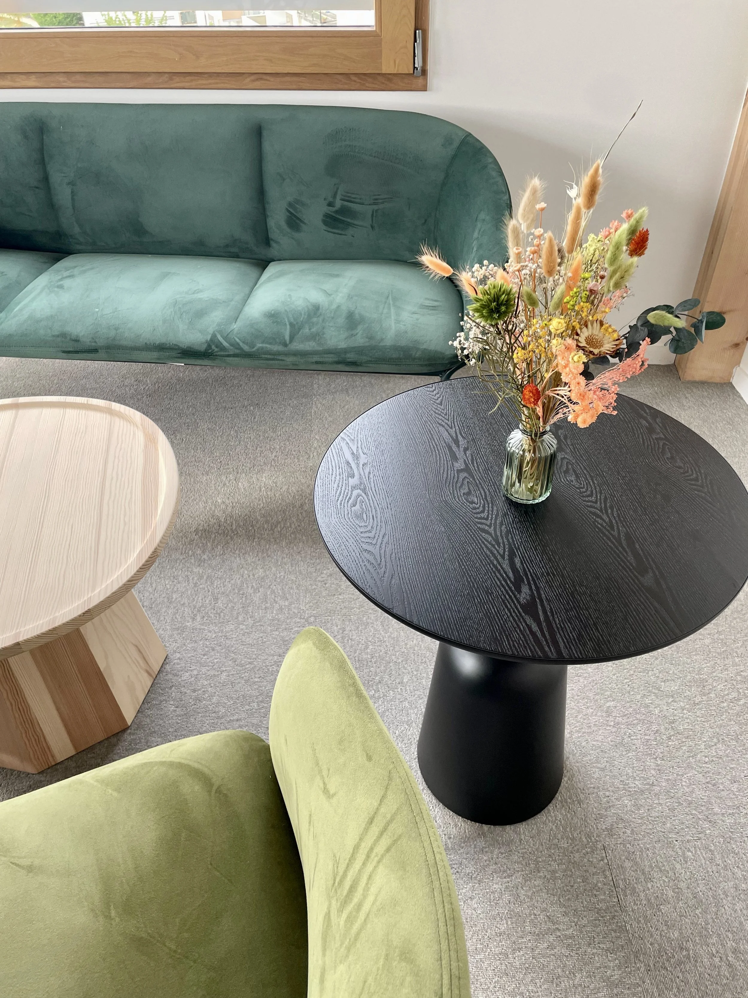 Une table noire avec un vase de fleurs séchées colorées, un canapé vert foncé et un fauteuil vert clair dans une pièce lumineuse.