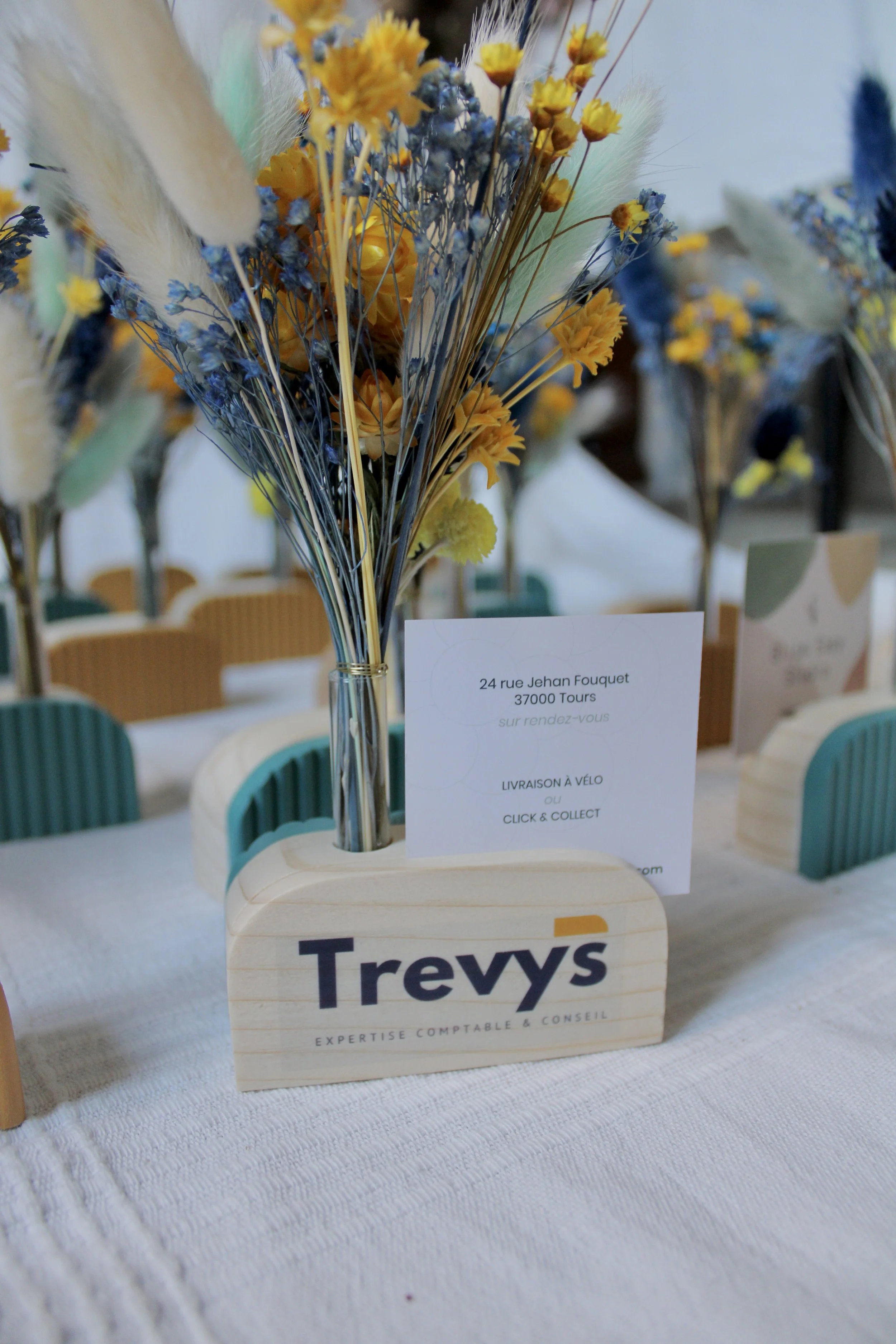 Une décoration florale avec un vase en bois ayant l'inscription 'Trevys' et une fiche d'information, le tout sur une table. Logo et fleurs séchées dans le vase.