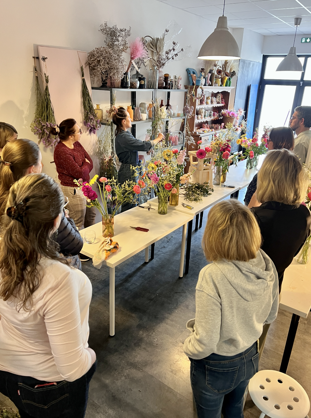 Groupe de personnes assises et debout dans un atelier de composition florale, avec une femme expliquant des techniques de décoration florale sur une table ornée de fleurs colorées.