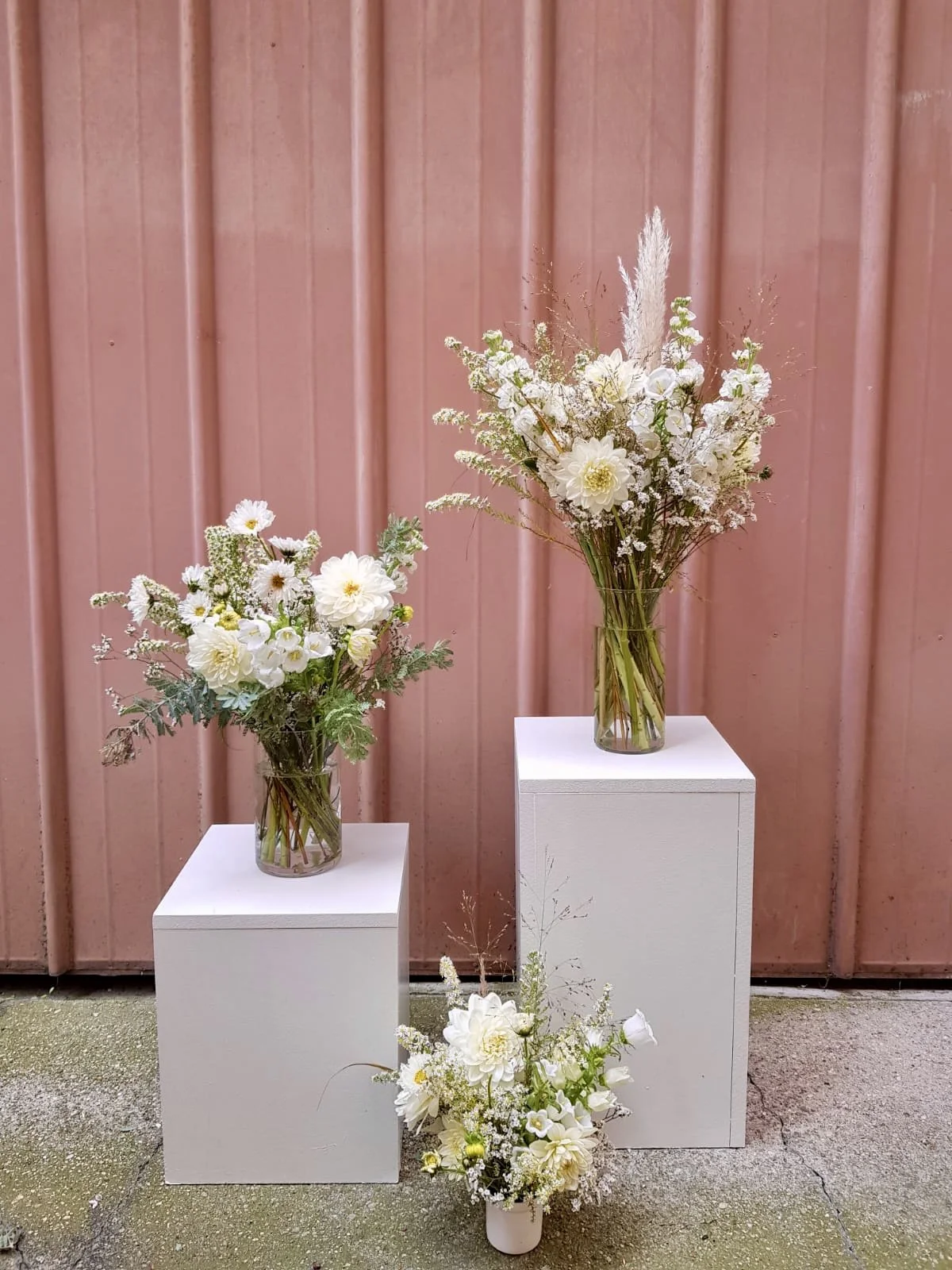 Trois arrangements floraux blancs dans des vases, exposés sur deux socles blancs et un sol, contre un mur en métal rose.