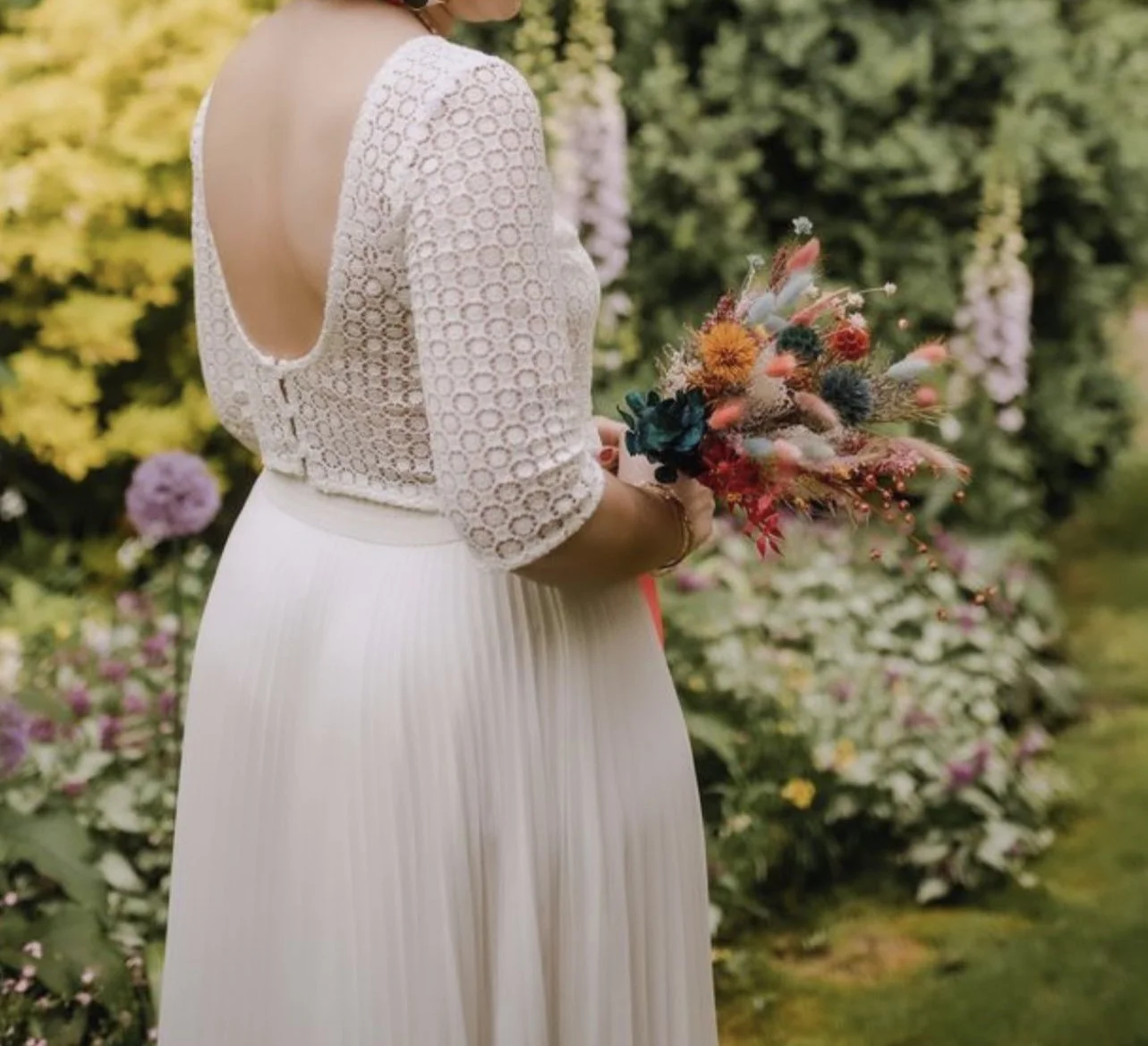 Femme portant une robe blanche avec un corsage en dentelle et un dos ouvert, tenant un bouquet de fleurs colorées dans un jardin fleuri.