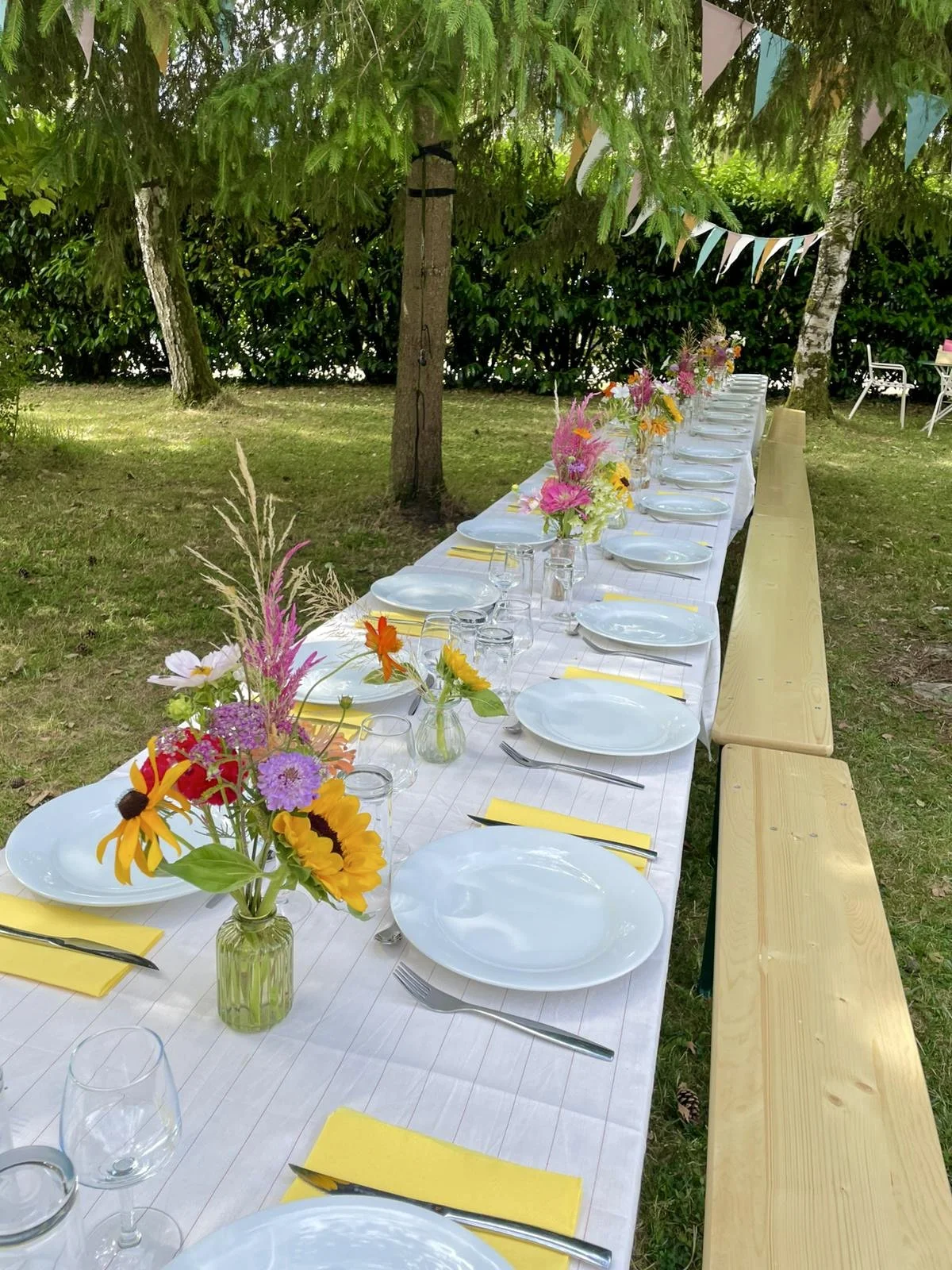 Une table de banquet décorée pour une fête en plein air, avec des assiettes blanches, des fourchettes, des verres, des bouquets de fleurs colorées dans des vases, et des banderoles triangulaires suspendues sous un arbre.