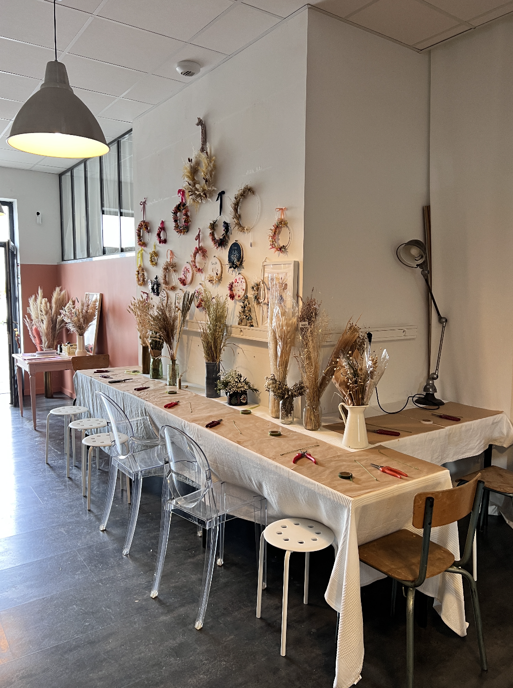 Décoration intérieure avec une table longue couverte d'une nappe blanche, ornée de bouquets de fleurs séchées, entourée de chaises en plastique transparentes et en bois, avec un mur décoré de couronnes florales.