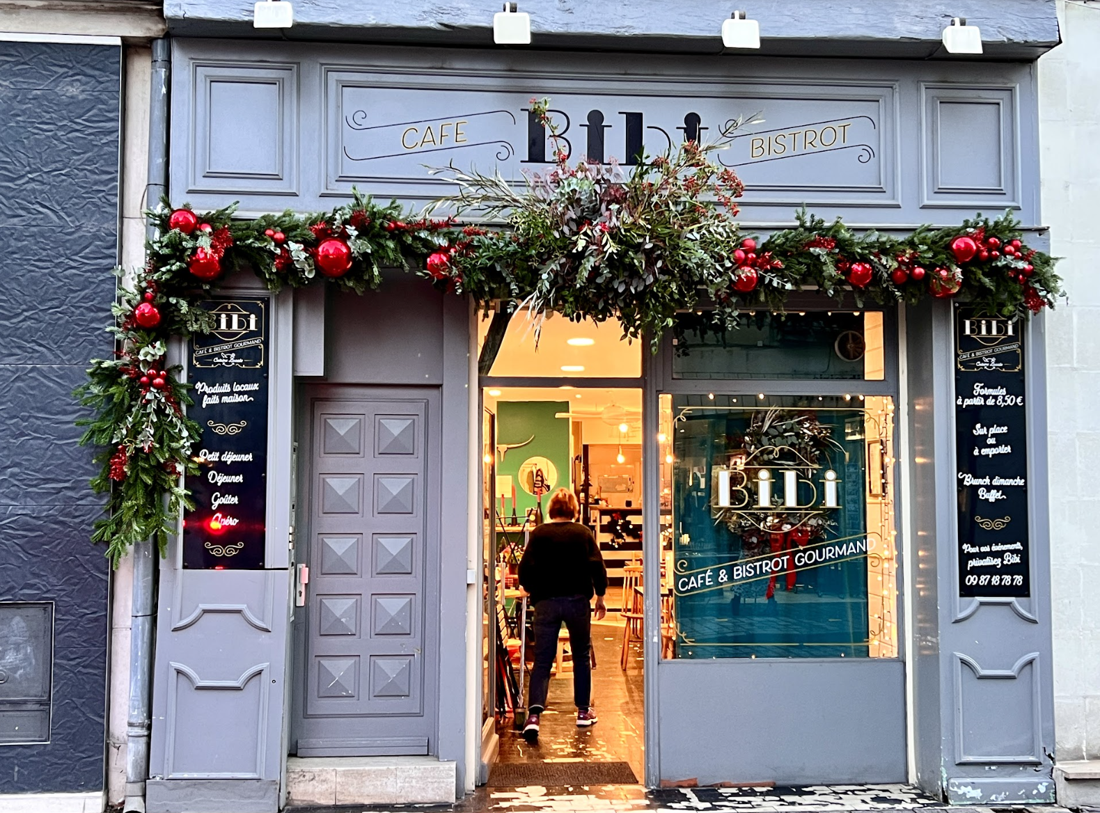 Une façade de café et bistrot décorée de guirlandes de sapin avec des boules rouges à Noël, une porte en bois gris et une grande vitre montrant l'intérieur chaleureux du restaurant.