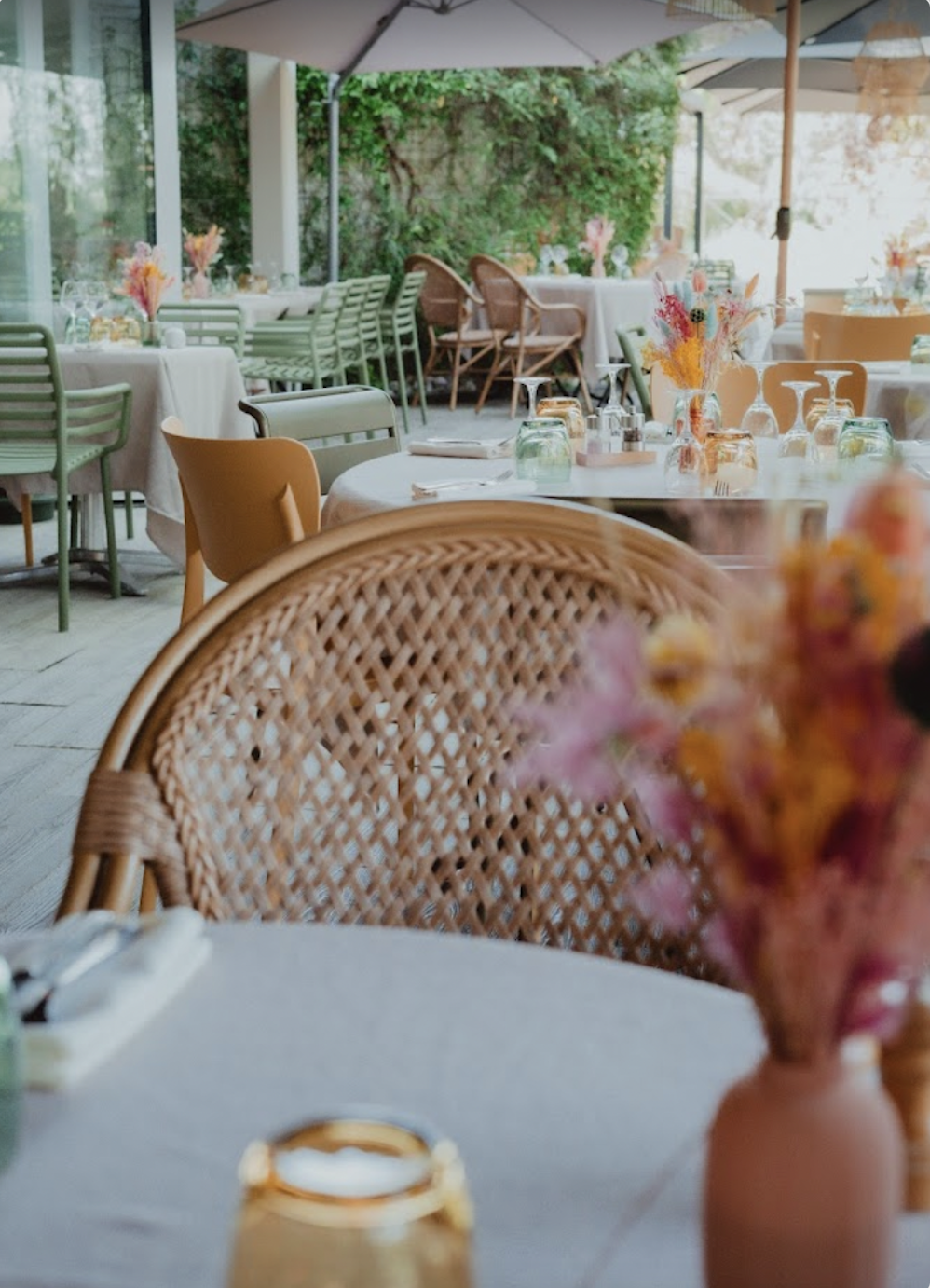 Salle à manger d'un restaurant ou d'une terrasse avec des tables ornées de vases avec des fleurs et des verres à vin, sous des parasols, avec un décor verdoyant à l'extérieur.