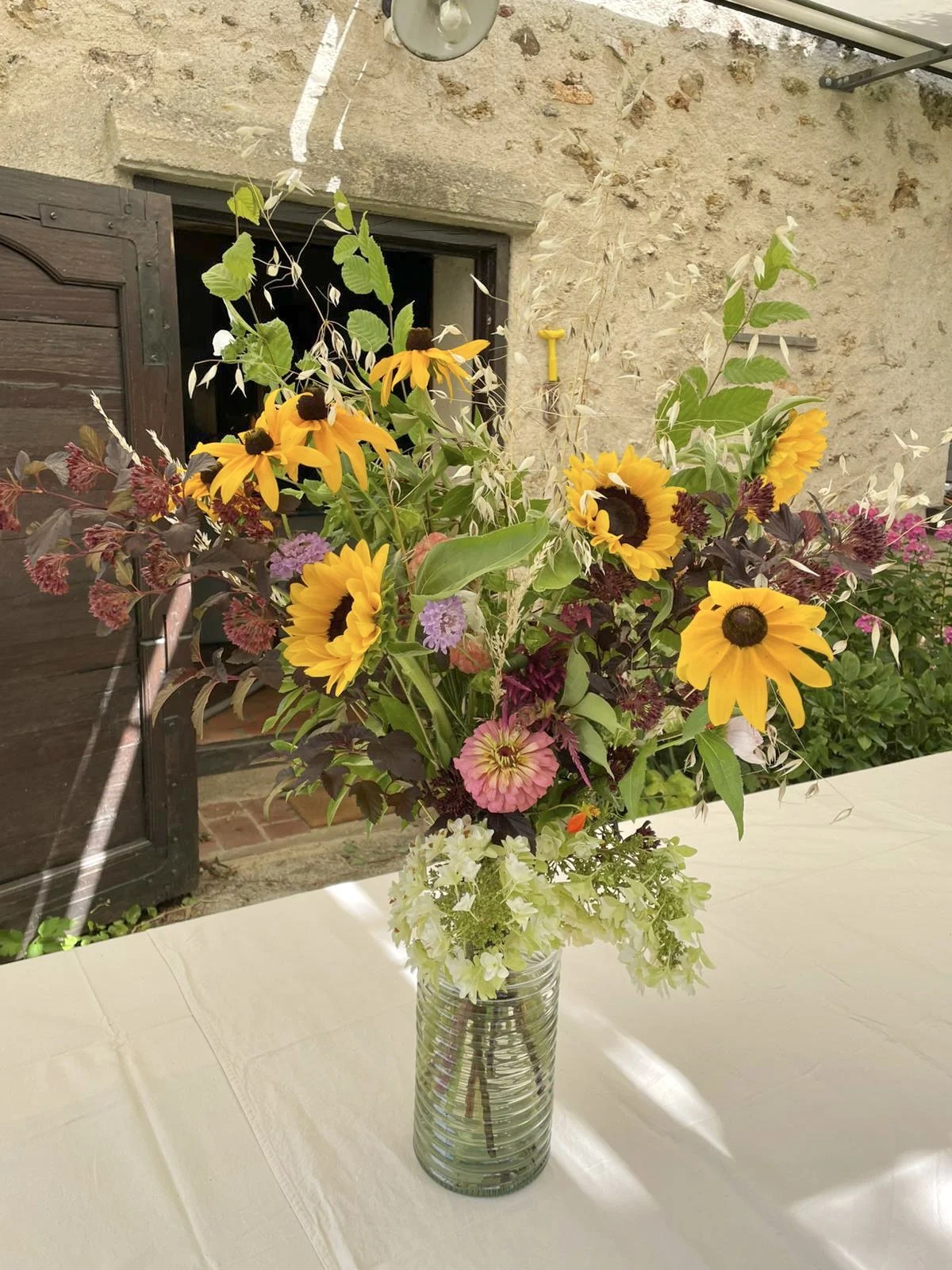 Un bouquet de fleurs colorées dans un vase en verre placé sur une table avec un fond de mur en pierre et une fenêtre.