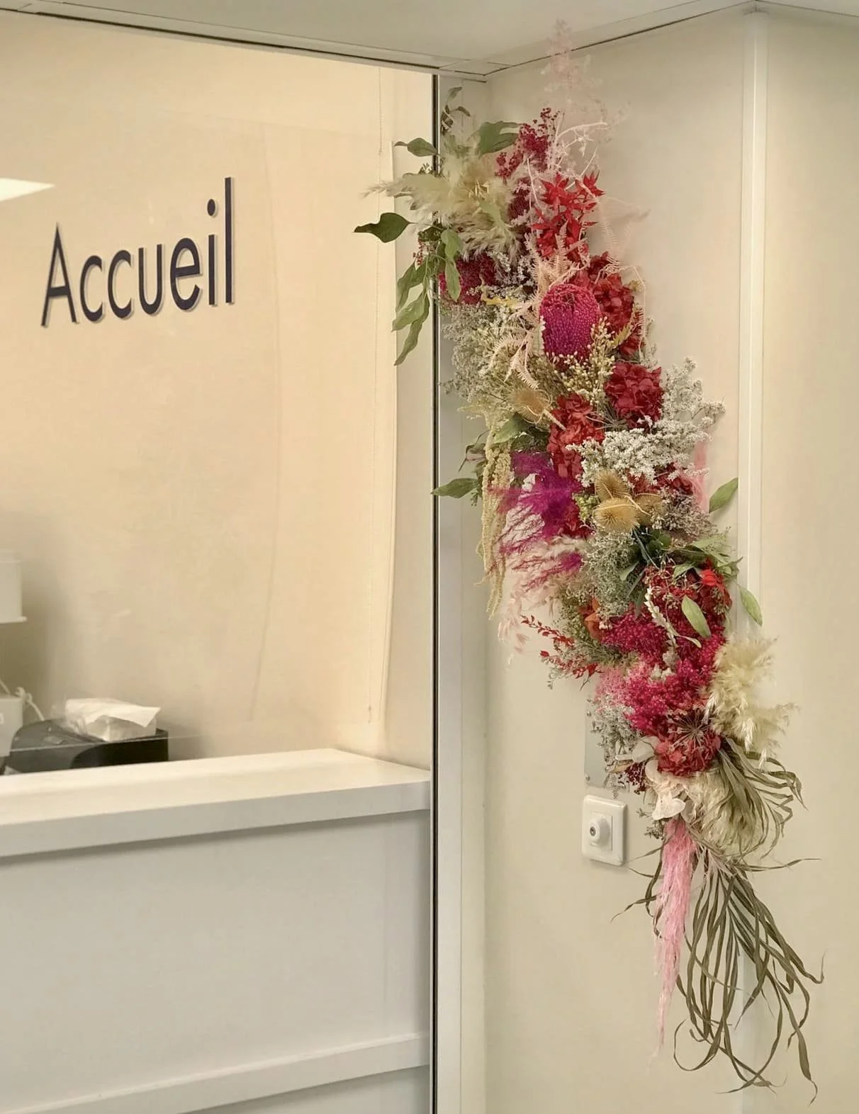 Décor floral suspendu avec différentes fleurs et feuillages colorés, situé à côté d'une porte d'entrée marquée 'Accueil' dans un intérieur.