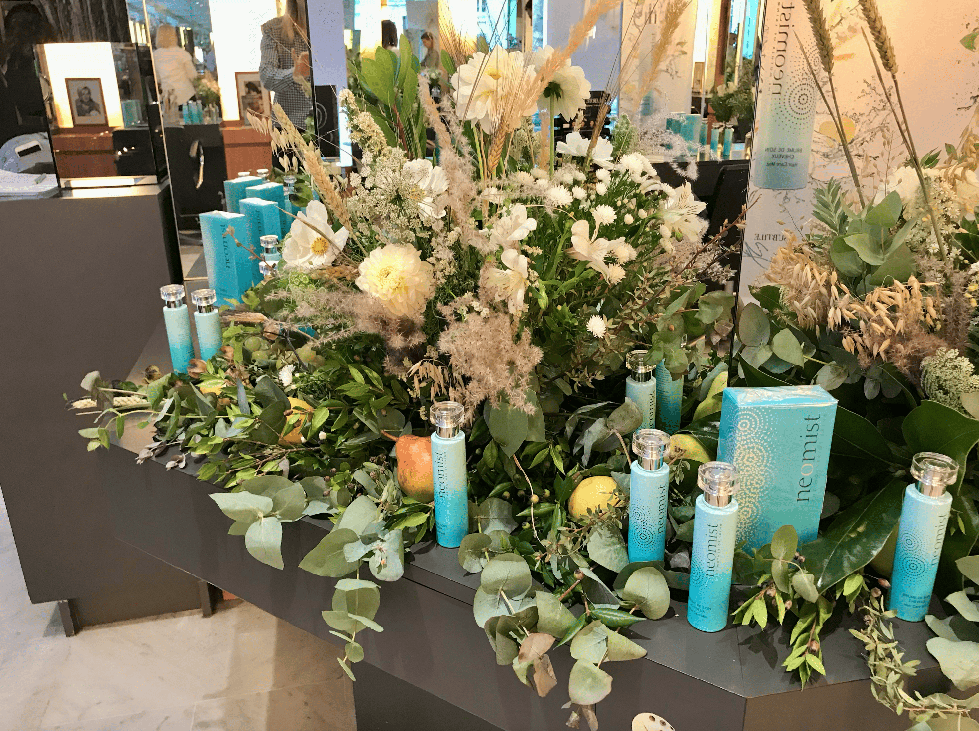Une grande composition florale composée de fleurs blanches et de verdure, entourée de plusieurs tubes et boîtes de produits de soin nommés 'neomist', sur un meuble sombre dans un environnement intérieur