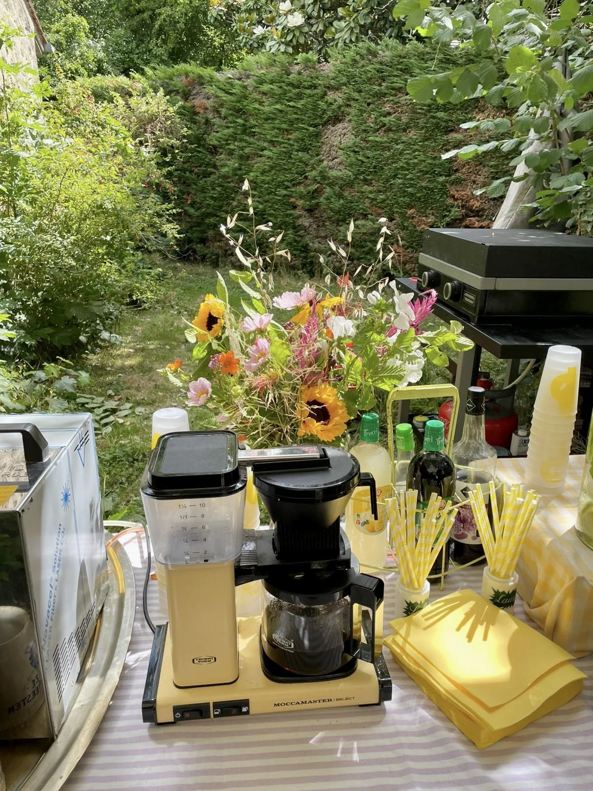 Une table avec une machine à café, des gobelets en papiers à rayures jaunes et blancs, des bouteilles de sirop, et un bouquet de fleurs colorées, en plein air dans un jardin avec des arbres et des buissons en arrière-plan.