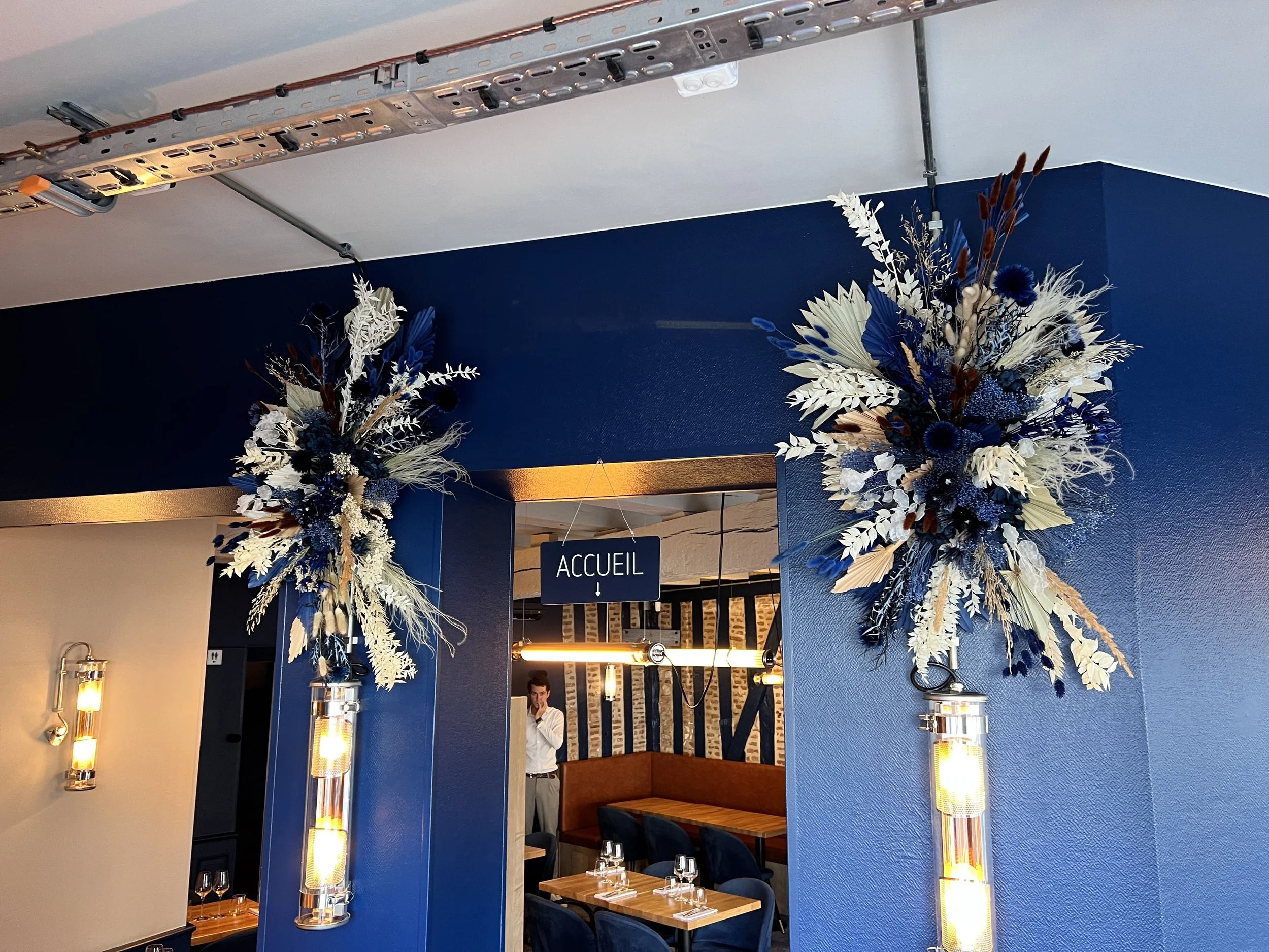 Décor de restaurant avec des arrangements floraux bleus et blancs épaisses sur les murs, un panneau 'Accueil' au plafond, tables prête à accueillir des clients, et un homme en tenue formelle en arrière-plan.