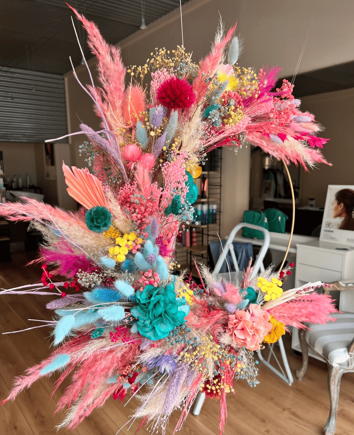 Un arrangement floral coloré avec des fleurs séchées dans des tons rose, bleu, jaune, violet et turquoise, placé sur une surface en bois dans une pièce intérieure.