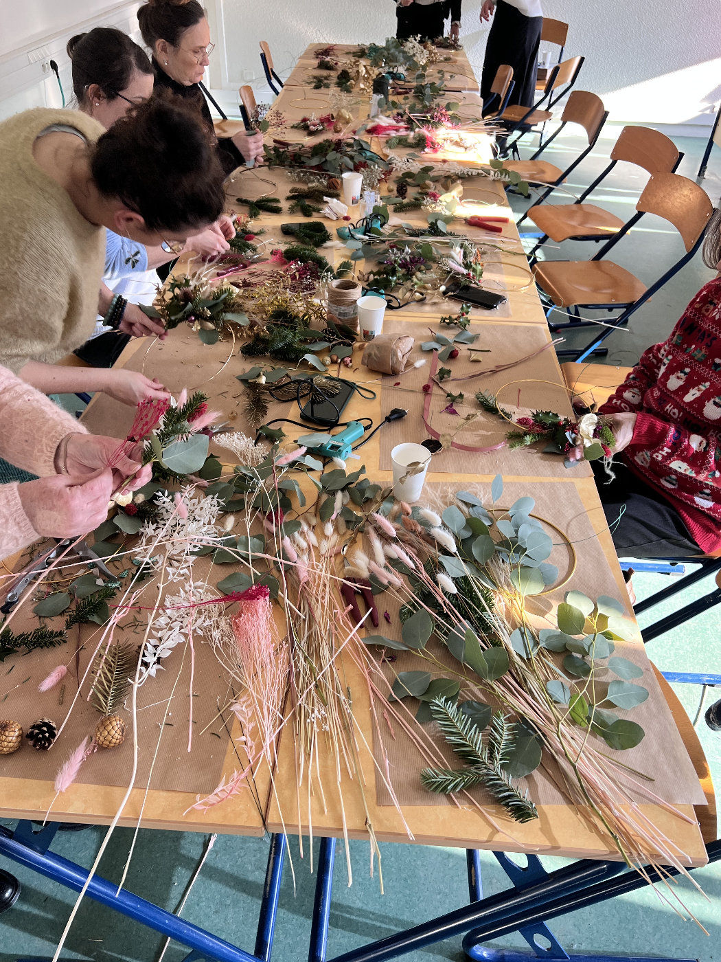 Groupe de personnes réunies autour d'une grande table en train de créer des décorations de Noël avec des branches de sapin, des feuilles, des rubans, et d'autres ornements.