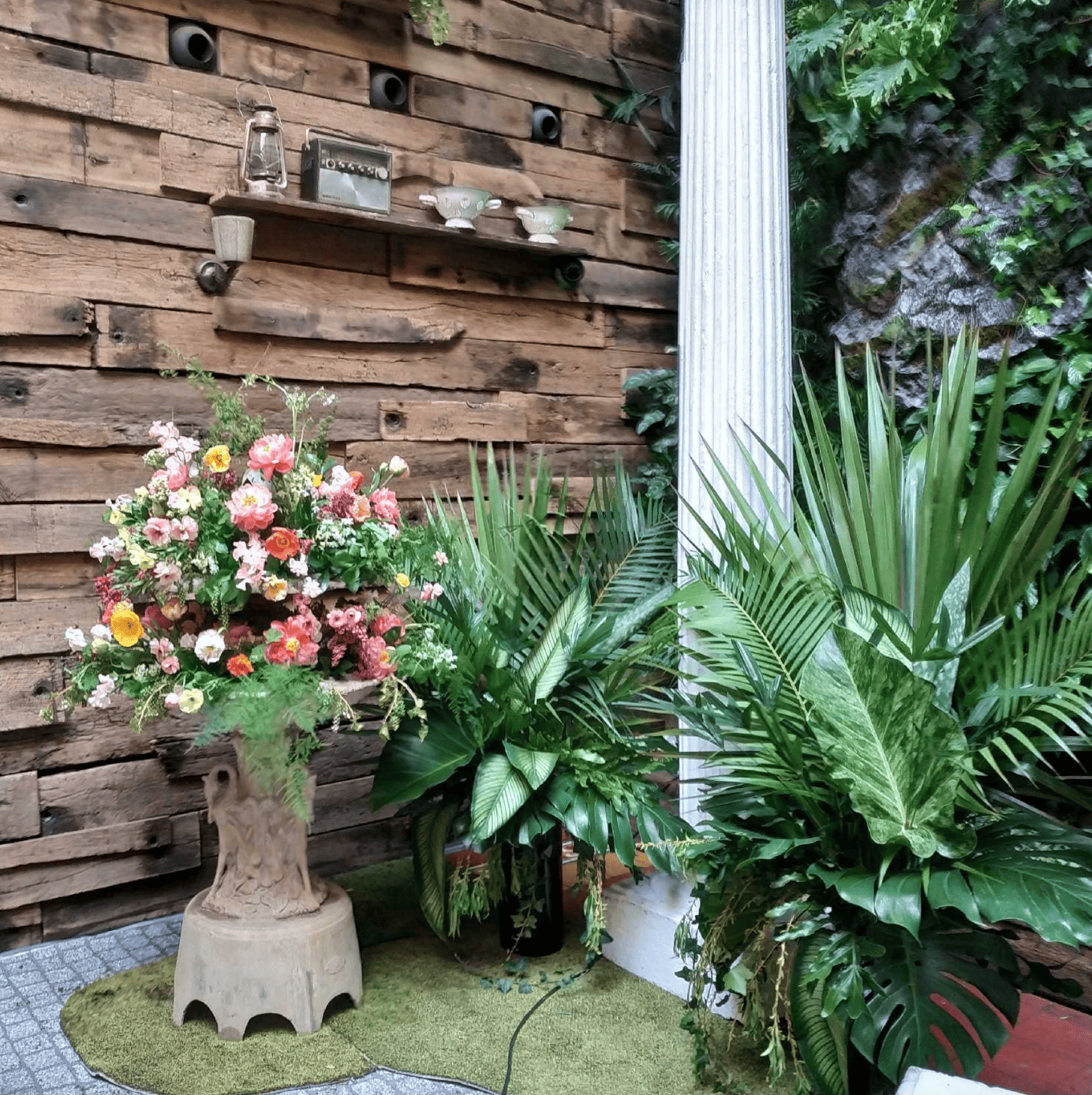 Un coin de jardin avec un mur en bois, une colonne en plastique, des plantes vertes, un arrangement floral dans une poterie et des étagères métalliques avec des objets décoratifs.