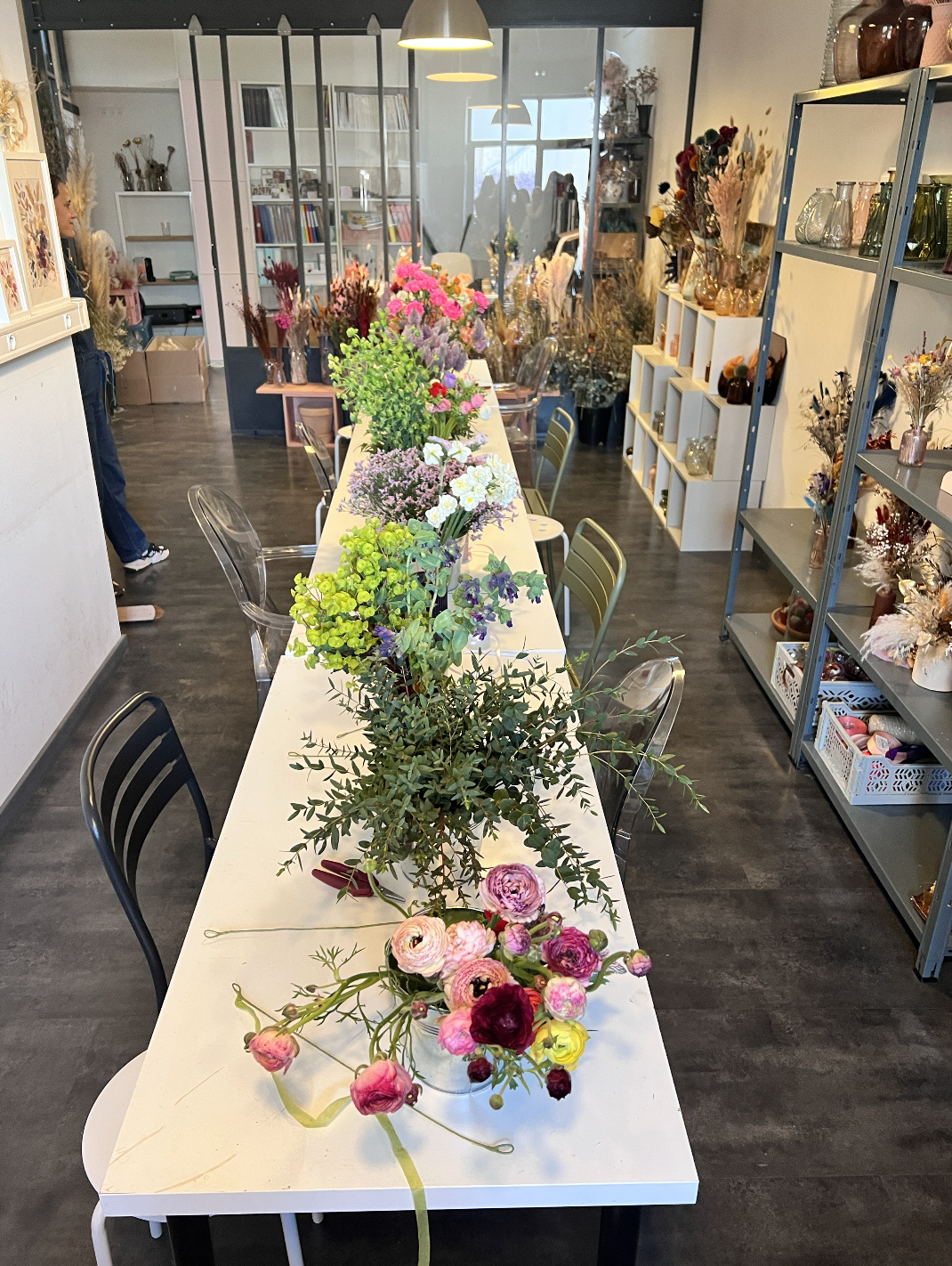 Une salle avec une longue table en blanc décorée de plusieurs arrangements floraux colorés, entourée de chaises transparentes et vertes, avec des étagères remplies de vases et décorations à l'arrière.