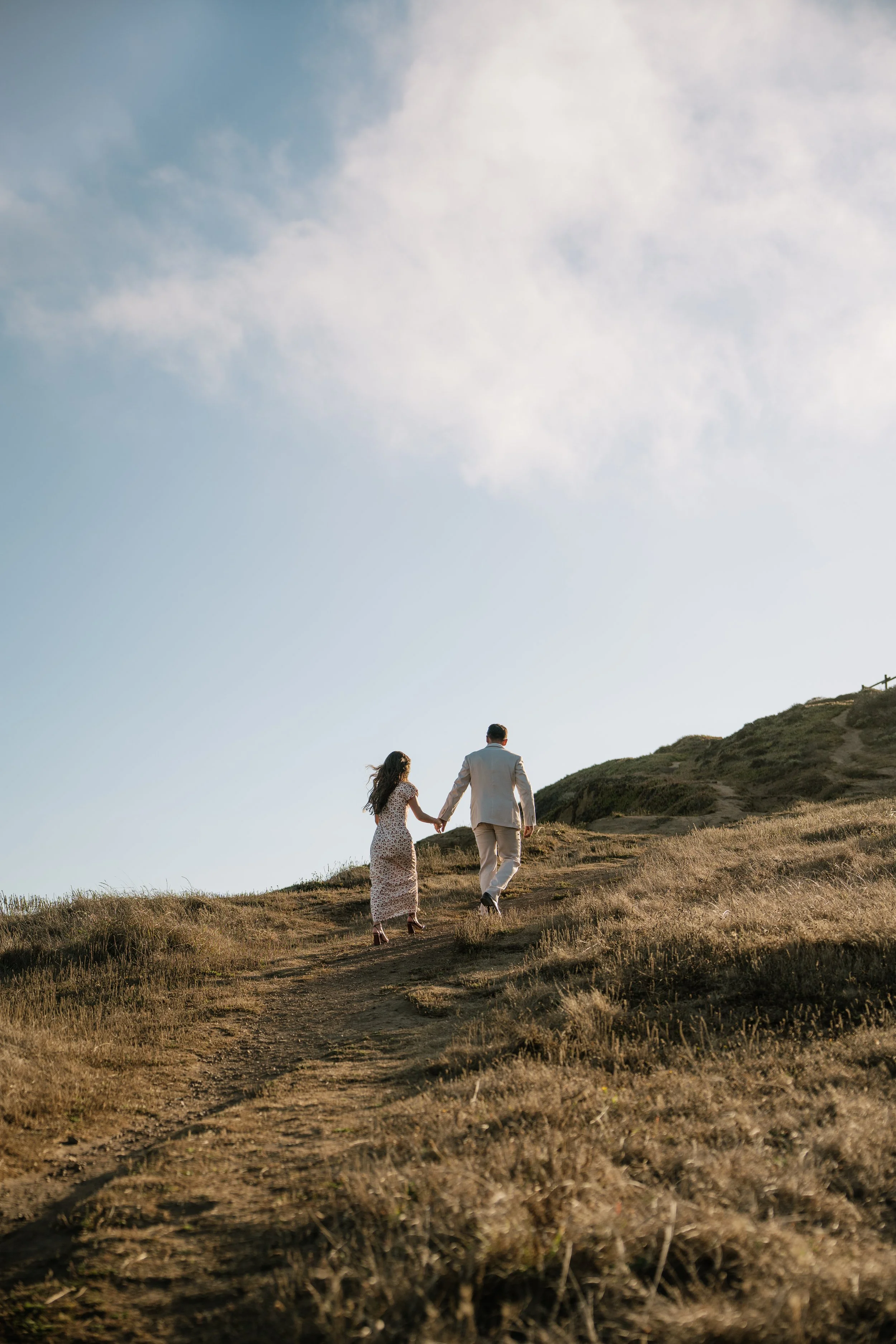 A engaged man and woman holding hands and walking up a grassy hill during sunset in San Fransisco, California.