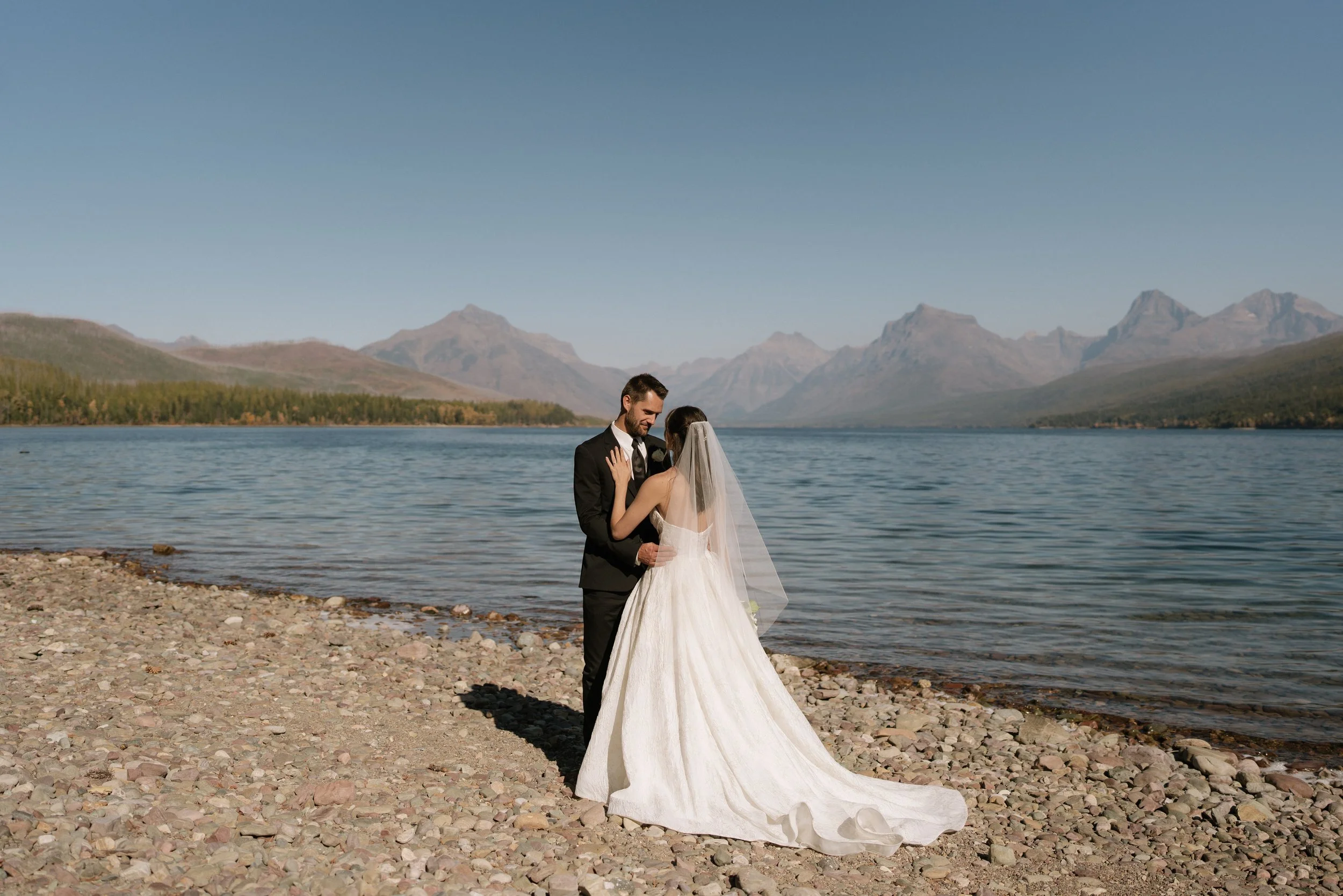 A bride and groom standing on a rocky shore by Lake McDonald, embracing, with the Glacier National Park mountains in the background under a clear blue sky.