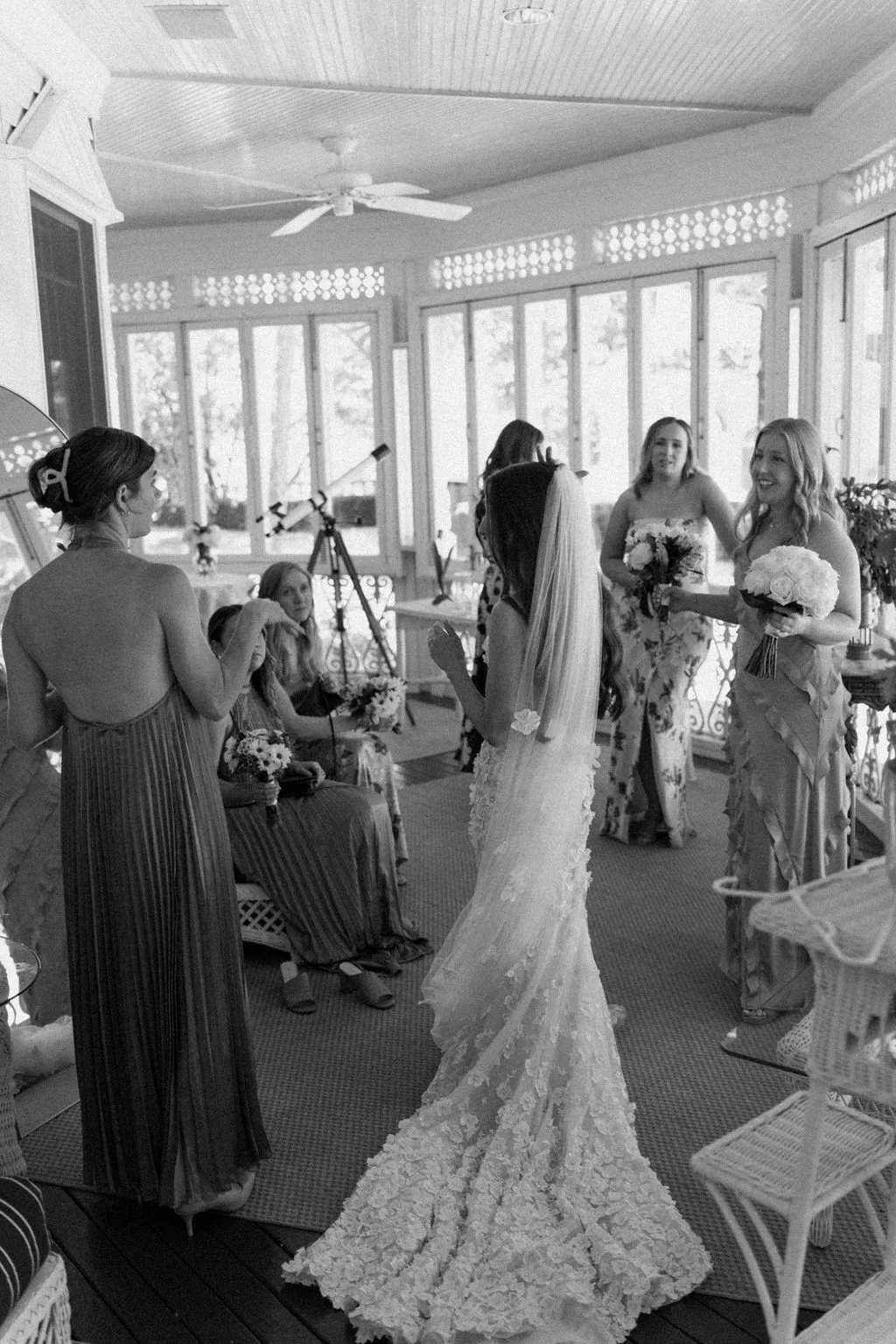 A black and white photo of a bride getting ready for her wedding featuring a bride in a lace wedding gown and veil talking to her bridesmaids, who are holding bouquets and wearing floral dresses. Decor and telescopes are visible in the background.