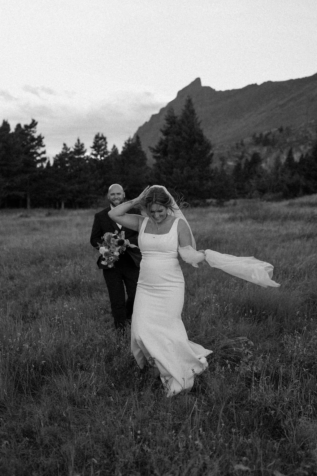 A bride and groom walking through a field with mountains and trees in the background in Glacier National Park, holding flowers, on a windy day. The photo is in black and white.