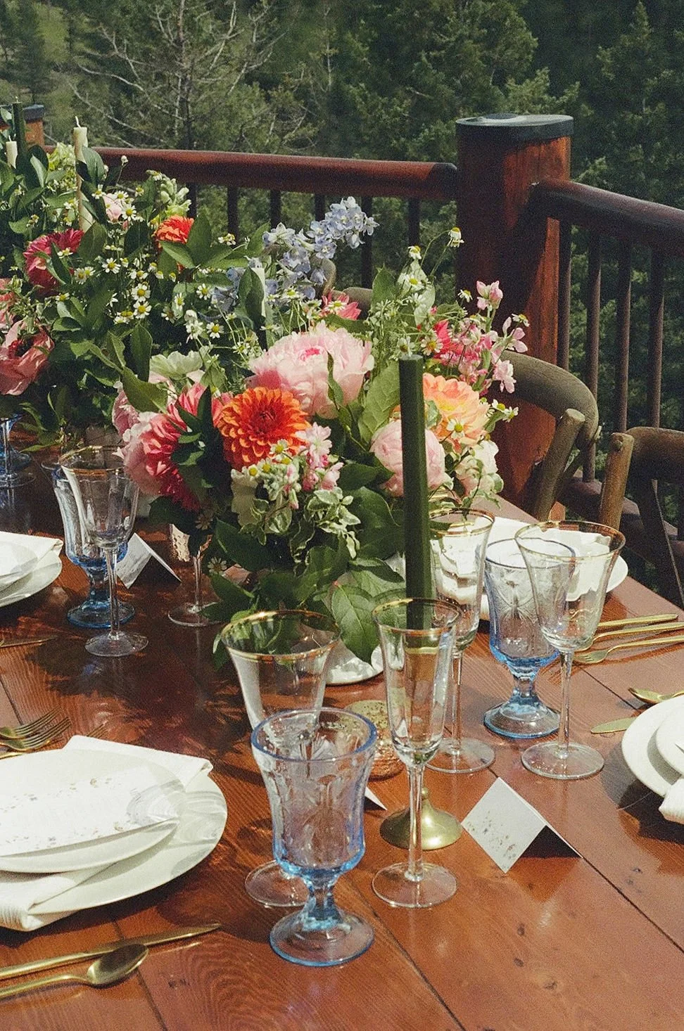 A film photo of a wooden outdoor dining table set with white plates, gold utensils, and a large colorful floral centerpiece, surrounded by chairs, with a scenic forest view in the background near Flathead Lake in Montana.