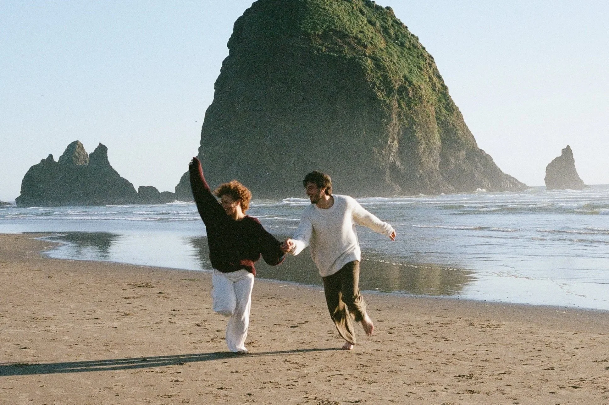A couple smiling and running on Cannon Beach holding hands and smiling.