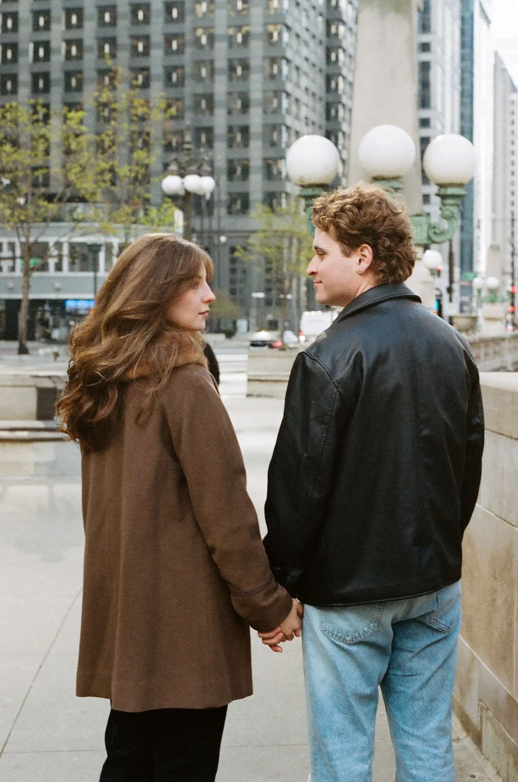 A film photo of a couple holding hands, standing outdoors on a city street in Chicago with tall buildings and vintage street lamps in the background, facing each other.