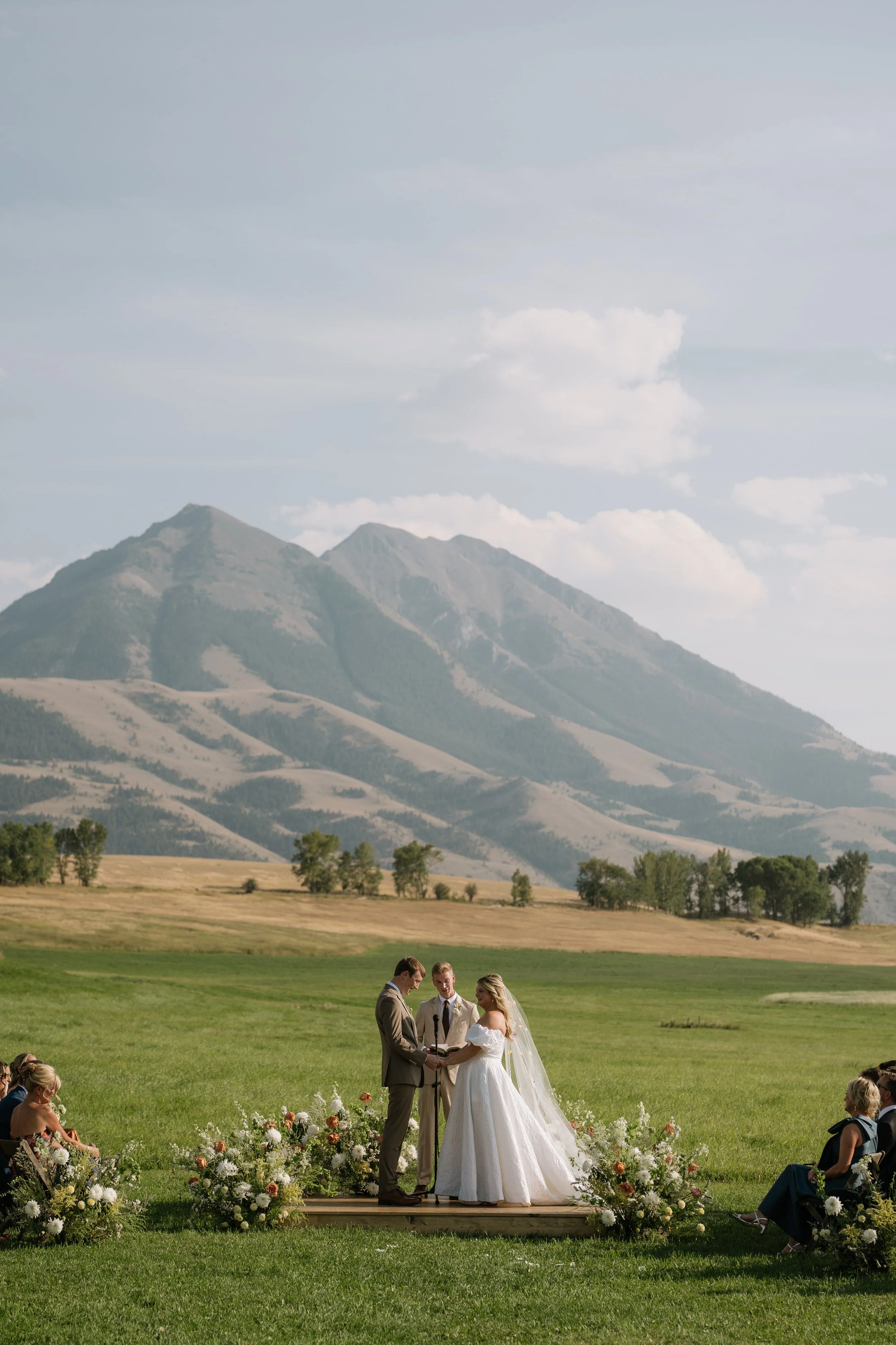 A wedding ceremony taking place at Sage Lodge in Montana featuring a bride and groom surrounded by floral arrangements, with guests seated on either side of the ceremony.