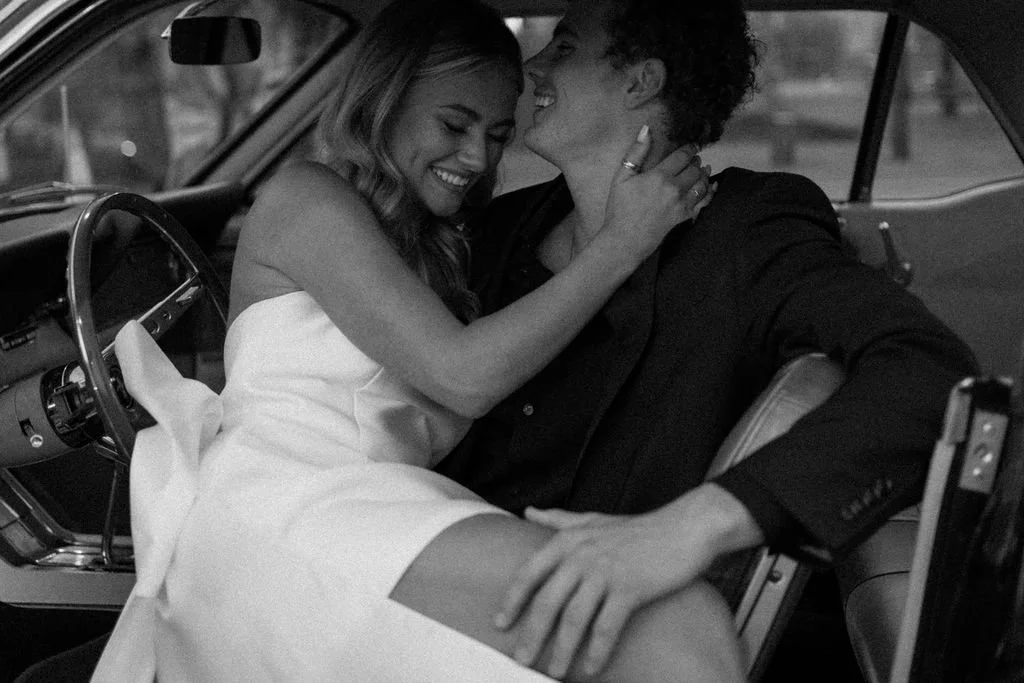 A man and women in a vintage car, one in a wedding dress and the other in a black suit, sharing a joyful and intimate moment in Nashville, Tennessee.