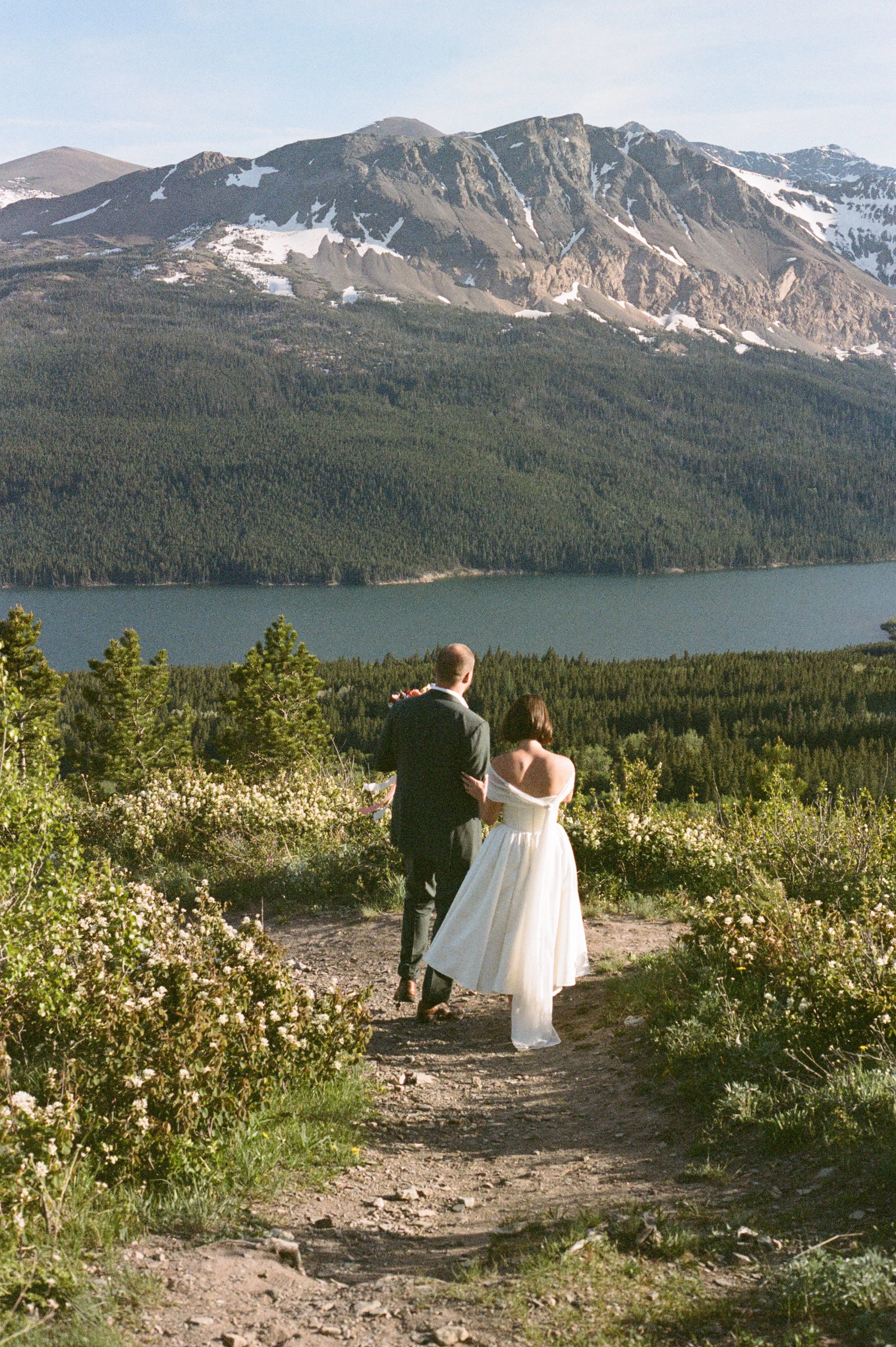 A couple dressed in wedding attire walking down a dirt trail with lush greenery and white flowers, overlooking lower Saint Mary Lake with mountains in the background near Glacier National Park.