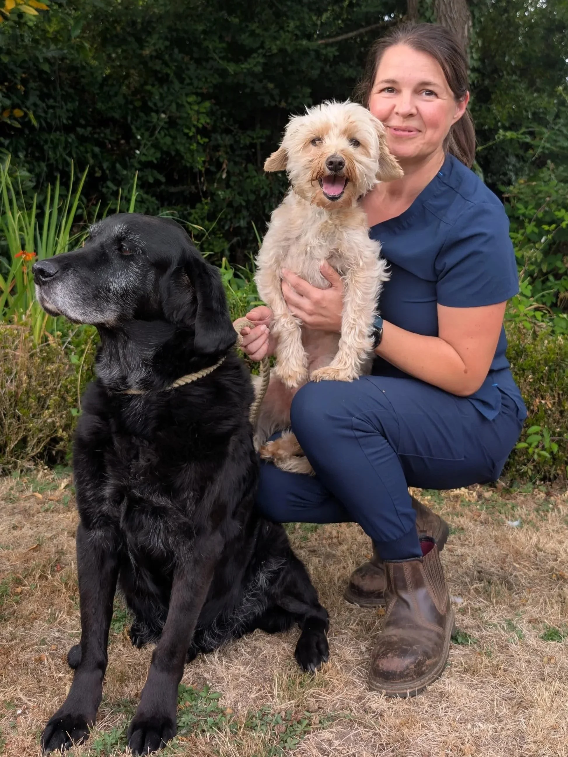 A woman in blue scrubs kneeling outdoors next to a black dog and a small tan and cream-colored dog, with trees and bushes in the background.