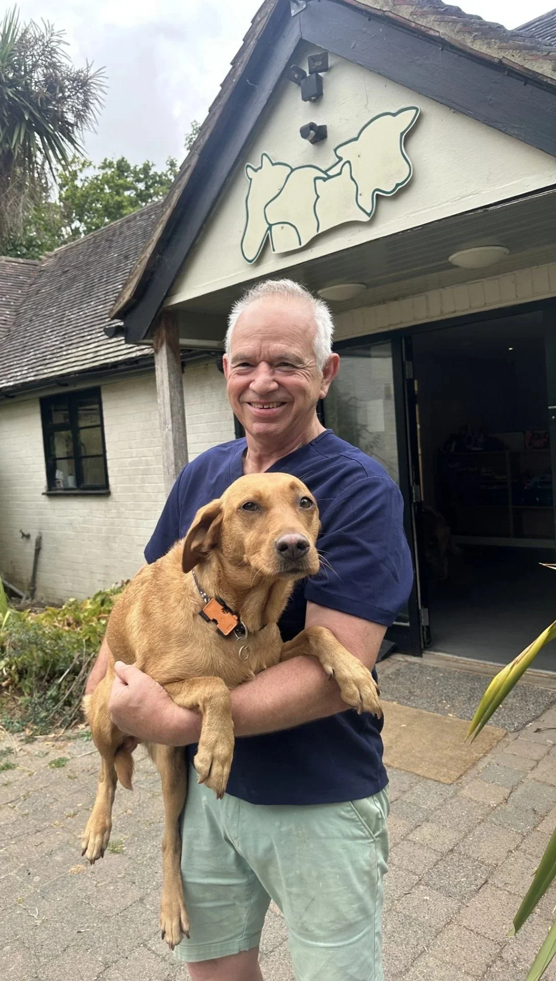 A man with gray hair smiling and holding a brown dog with floppy ears outside a building with a dog-themed sign above the entrance.