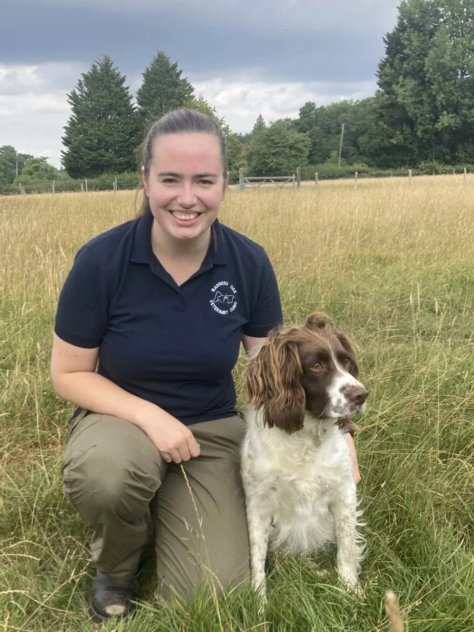 A woman kneeling in a field with a brown and white Springer Spaniel dog. She is wearing a navy polo shirt and beige pants, and smiling at the camera. The background has trees and a wooden fence under a cloudy sky.
