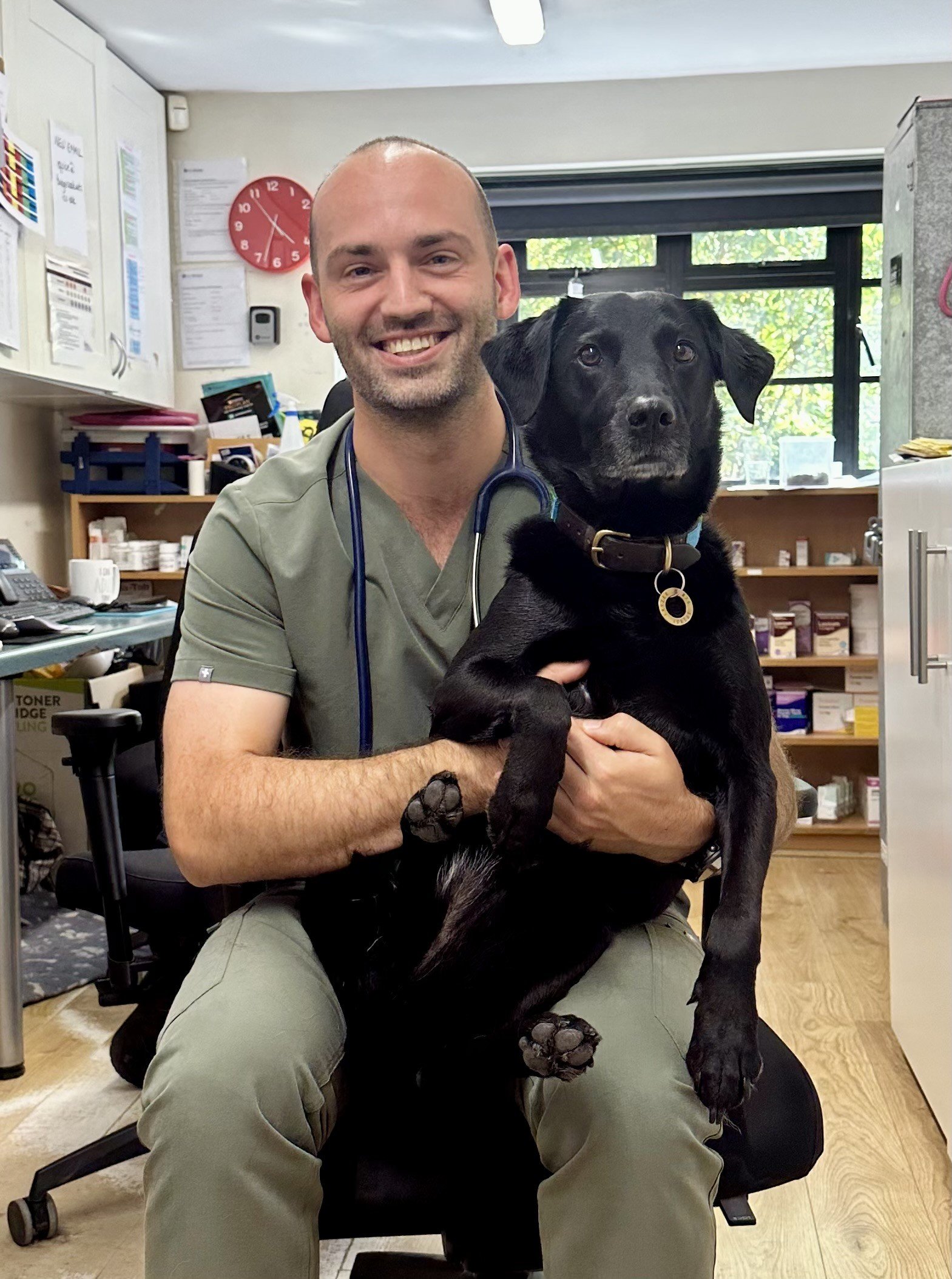 A man wearing scrubs and a stethoscope sitting on a chair in a veterinary clinic, holding a large black dog with floppy ears and a brown collar.
