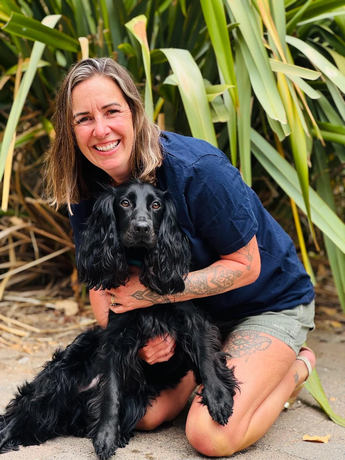 A smiling woman with tattoos on her arms and legs is kneeling outdoors, holding a black Cocker Spaniel dog with long ears and wavy fur. They are in front of tall green plants.