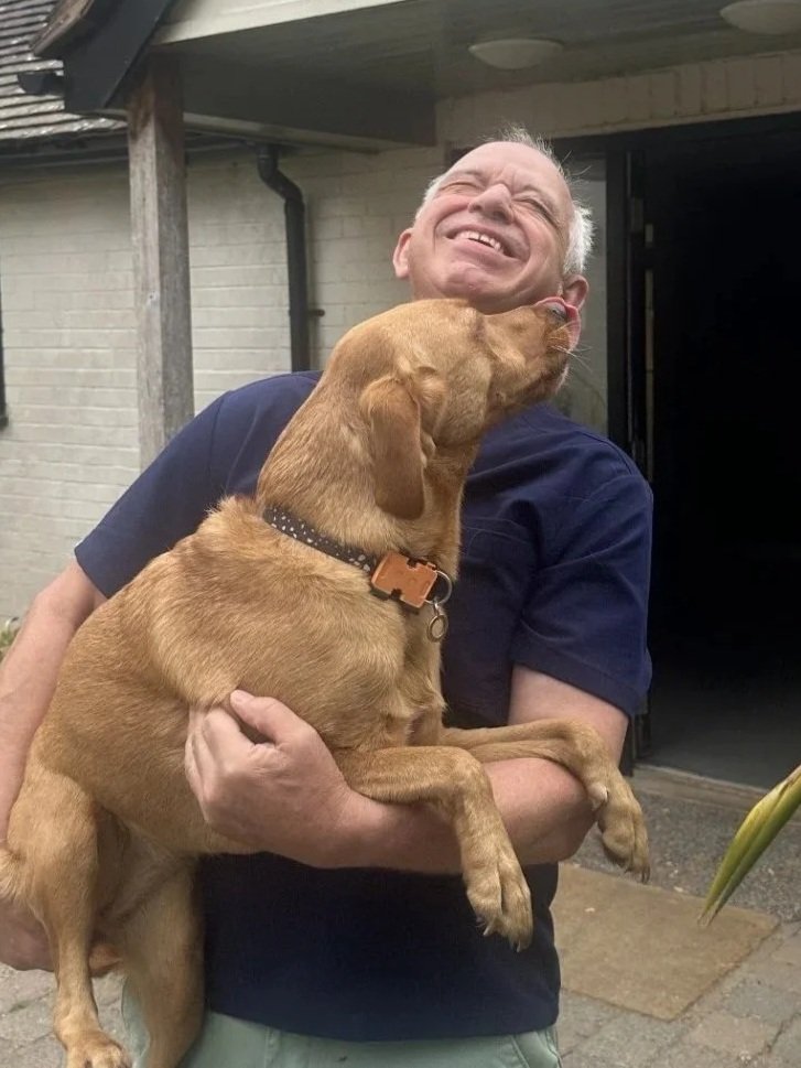 A man smiling and hugging a brown dog, which appears to be licking his face, outside Badgers Oak Veterinary Clinic