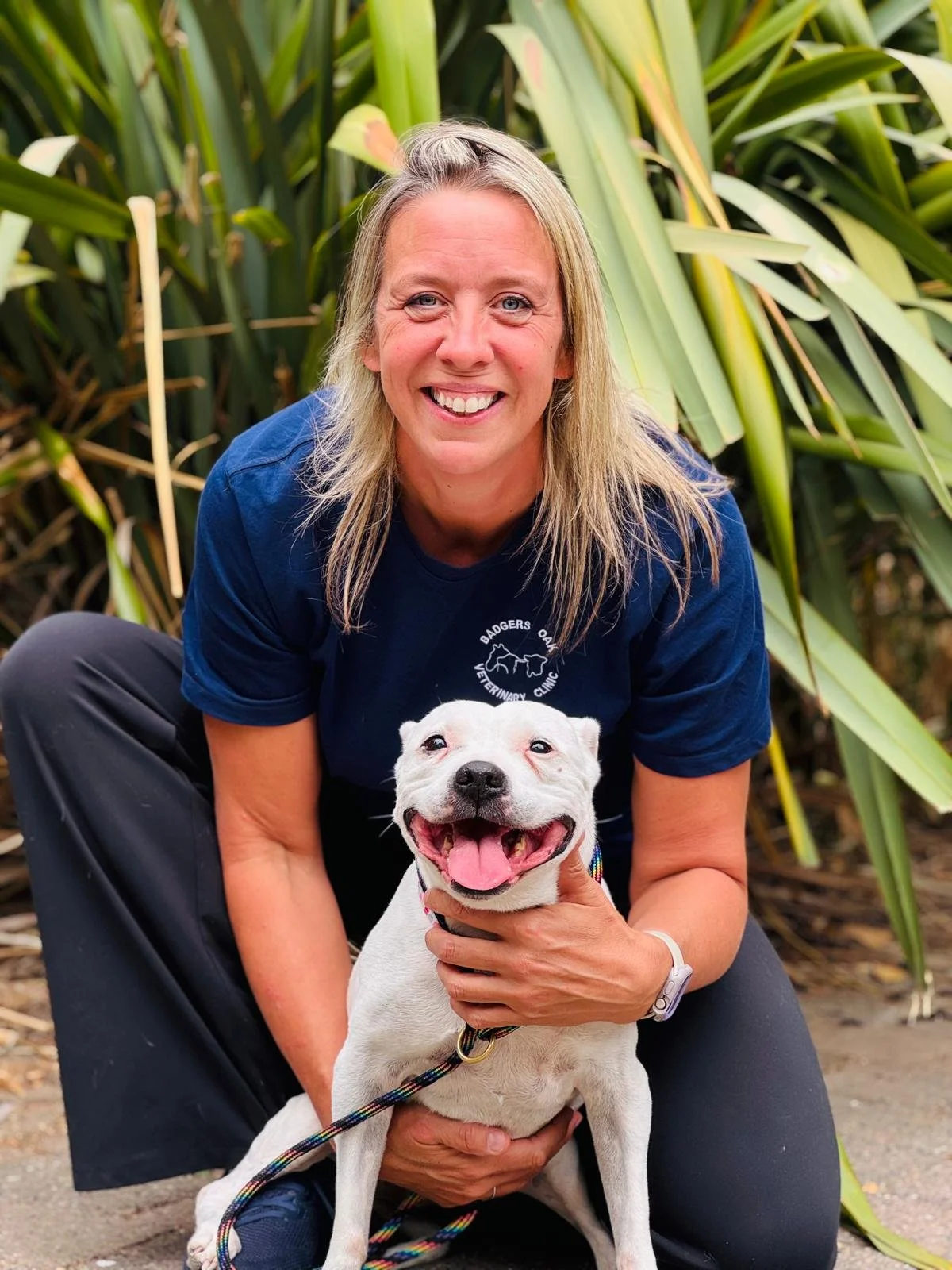 A woman with blonde hair smiling and crouching behind a happy white dog, with large green plants in the background.