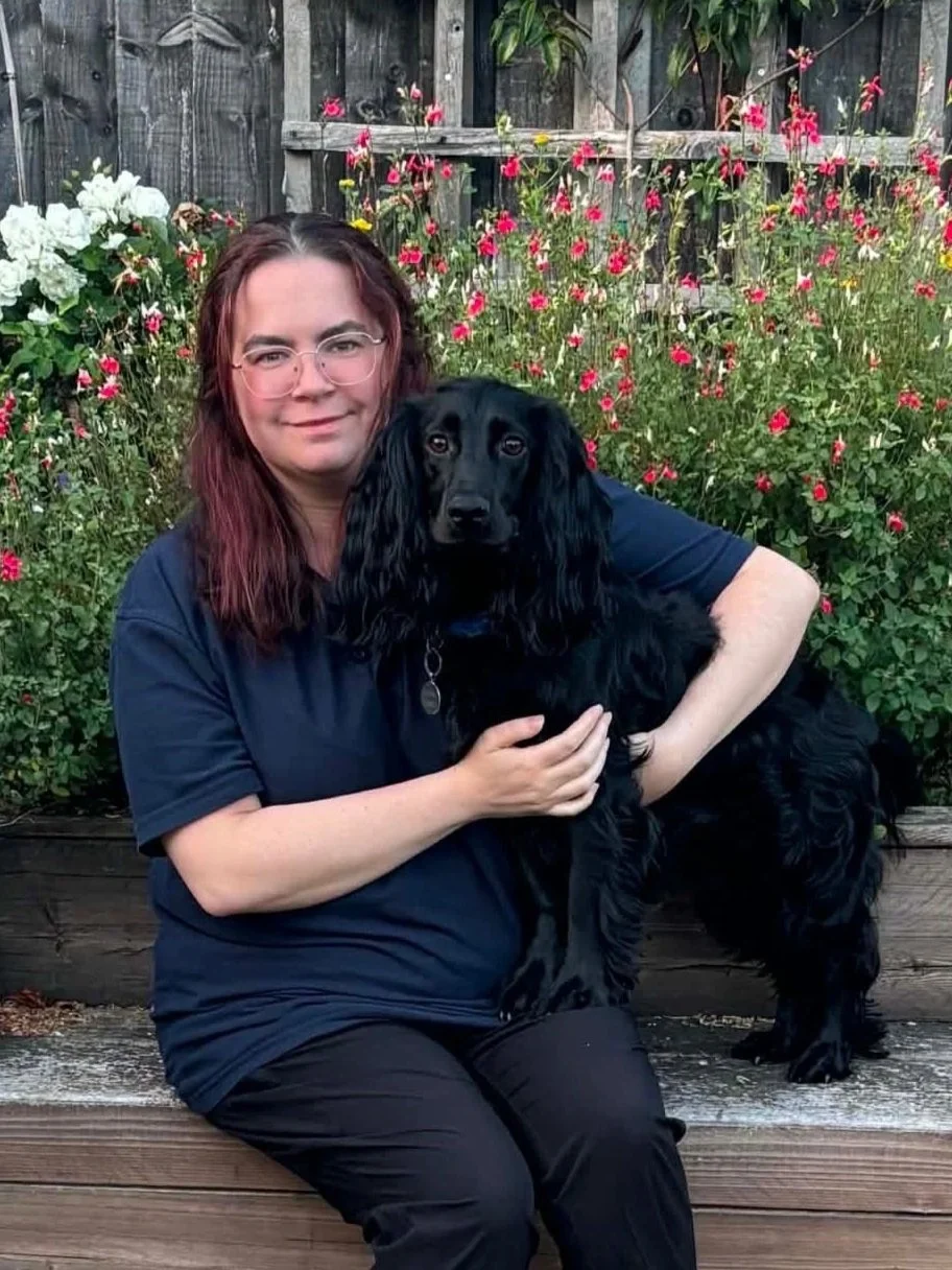 A woman with glasses and red hair sitting outside on wooden steps, holding a large black dog in front of a flower garden with pink, red, and white blooms.