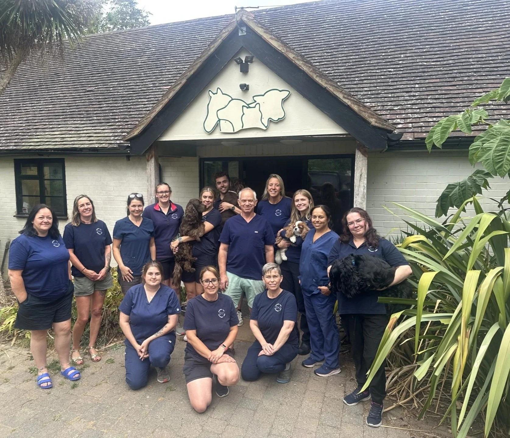 Group of people, both men and women, standing outside in front of a building with a dog-themed sign, all wearing matching dark blue scrubs, some holding dogs, at Badgers Oak Veterinary Clinic