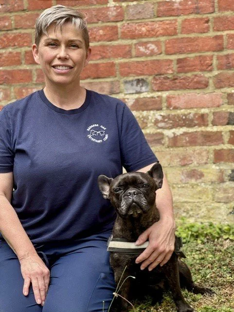 A woman with short gray hair smiling and sitting outdoors in front of a brick wall, holding a black French Bulldog wearing a harness.