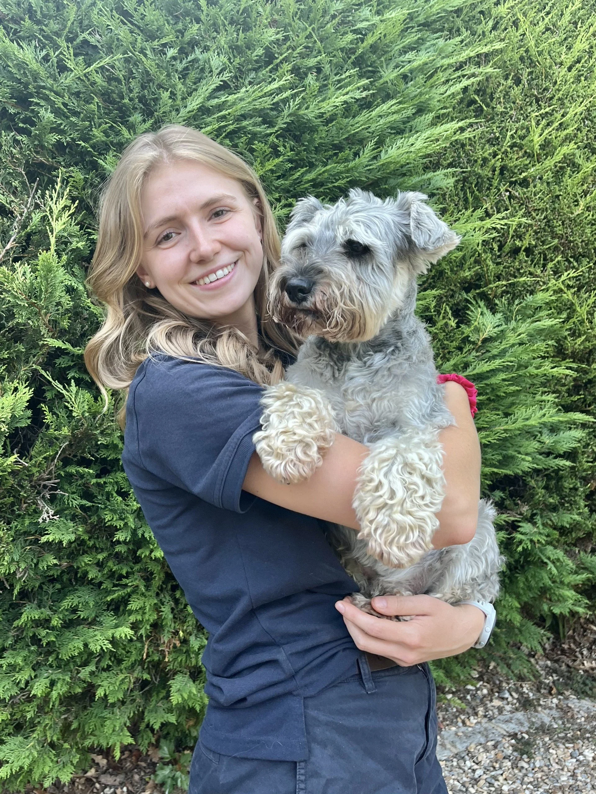 A young woman with blonde hair smiling and holding a gray and white dog with curly fur in front of lush green foliage.
