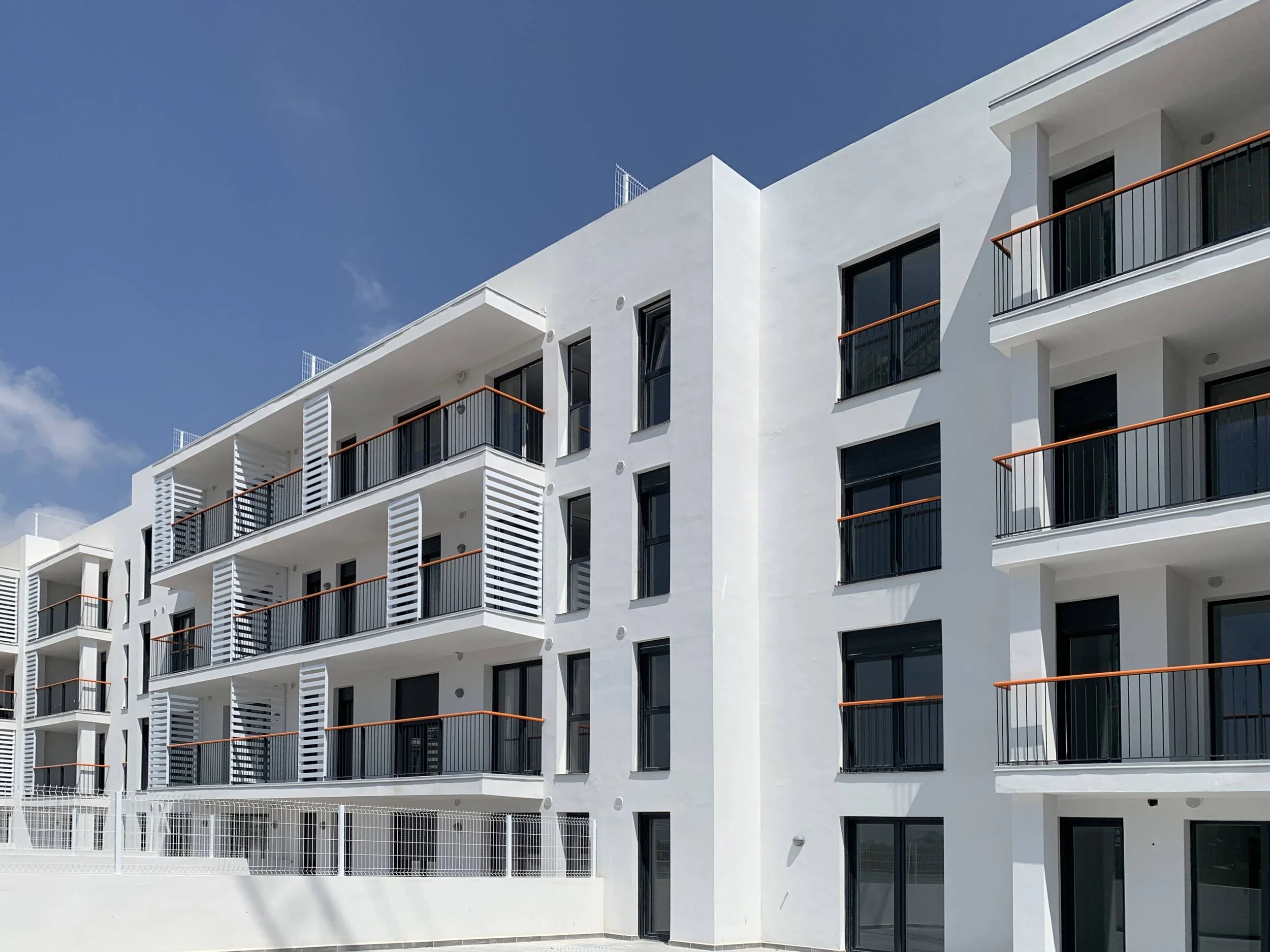Edificio moderno de apartamentos blancos con balcones con barandillas negras y detalles en madera, bajo un cielo azul.