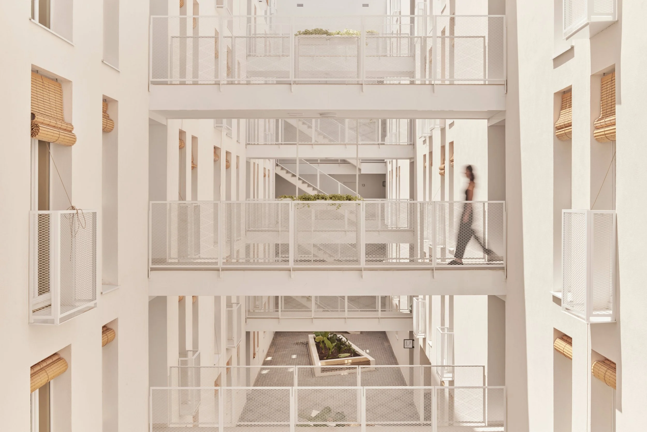 Interior de edificio de apartamentos con pasillos y balcones blancos, una mujer caminando por el pasillo en el nivel medio, plantas en un patio interior en la planta baja.
