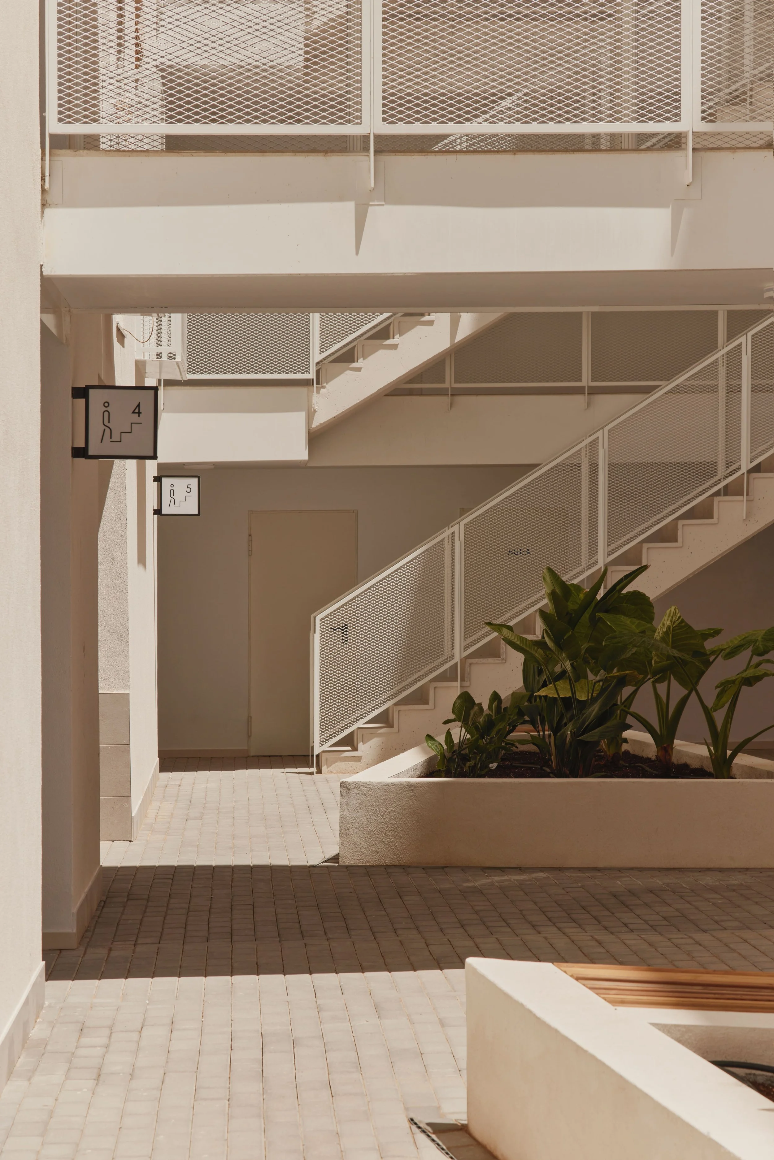Pasillo exterior de un edificio con escaleras y señalización de pisos en blanco y negro, plantas verdes en una jardinería y paredes de color crema.