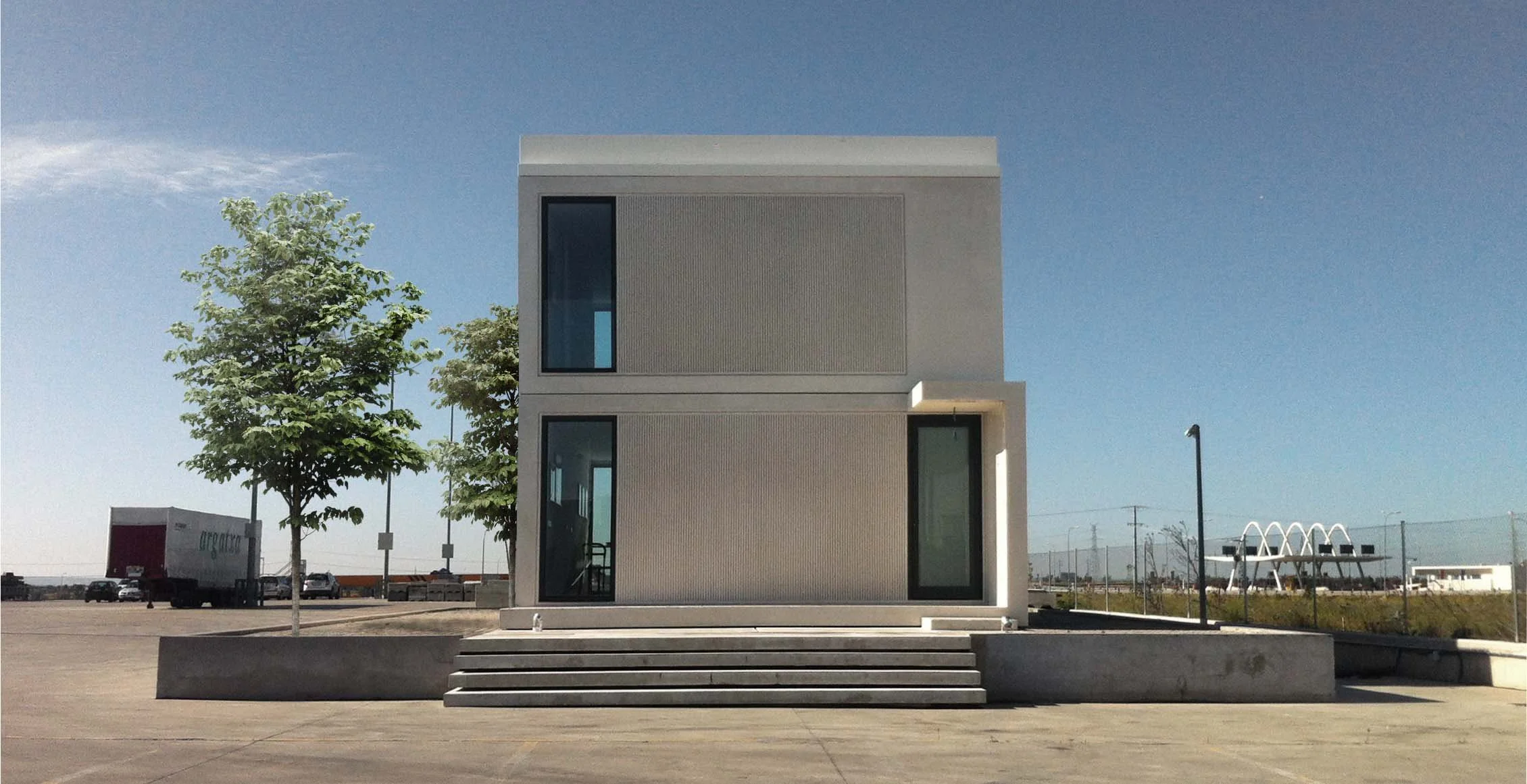 Edificio moderno de dos pisos con escaleras al frente, en un área abierta y despejada, con árboles y un cielo despejado.