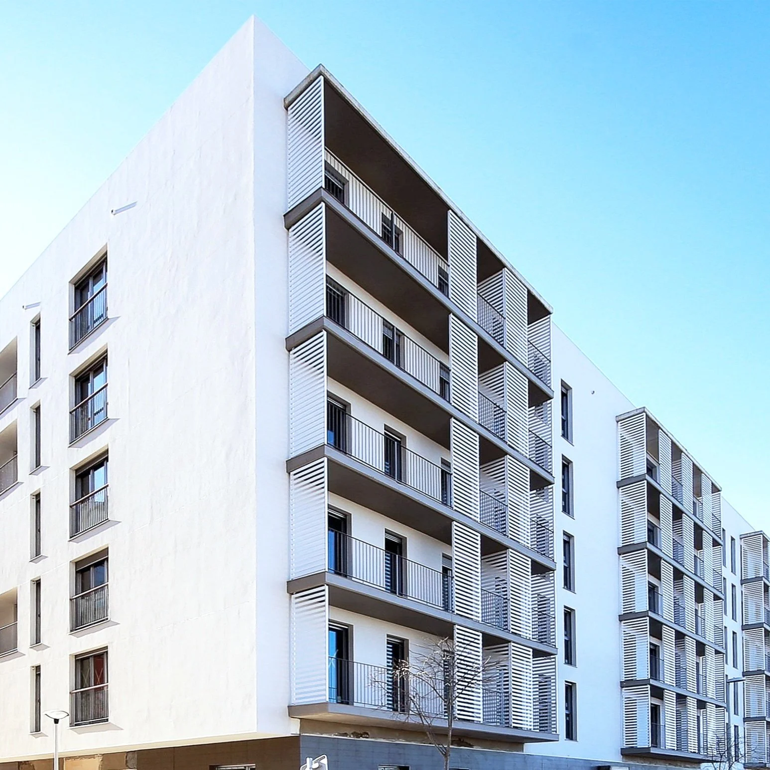 Edificio moderno de apartamentos con balcones y persianas blancas, bajo cielo azul.