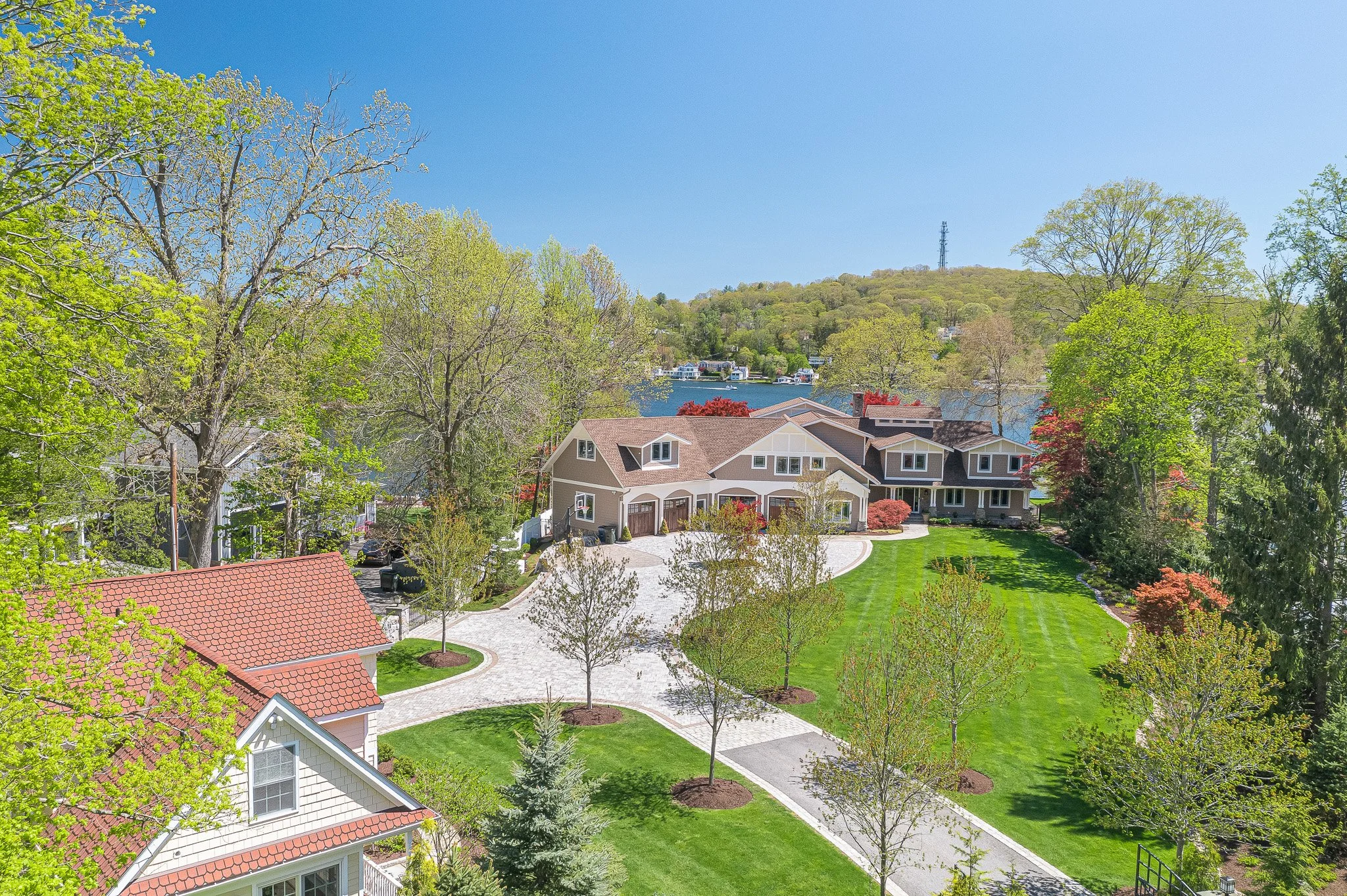 A large residential estate with a manicured lawn, trees, and a winding driveway leading to a house with multiple sections and large windows, near a lake with trees on the opposite shore under a clear blue sky.