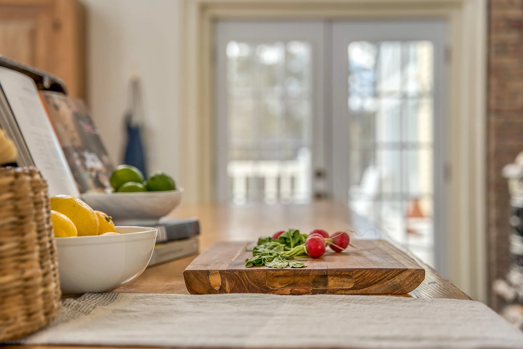 Fresh radishes and leafy greens on a wooden cutting board in a kitchen with bowls of lemons and limes nearby.