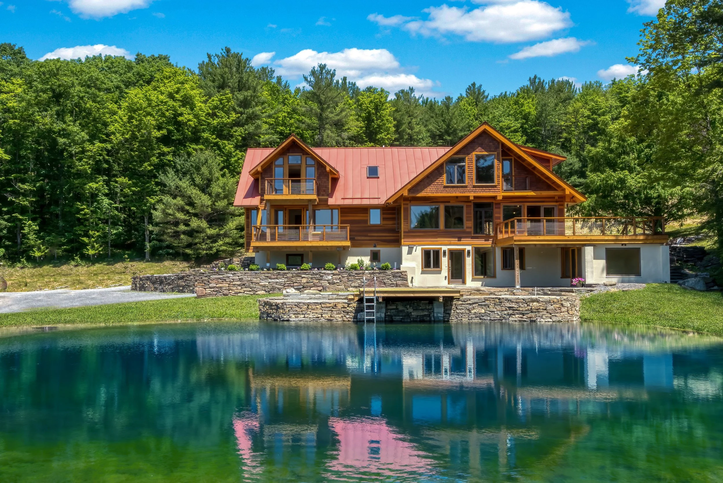 A large modern house with wooden and stone exterior faces a pond, surrounded by green trees and a blue sky with fluffy clouds.