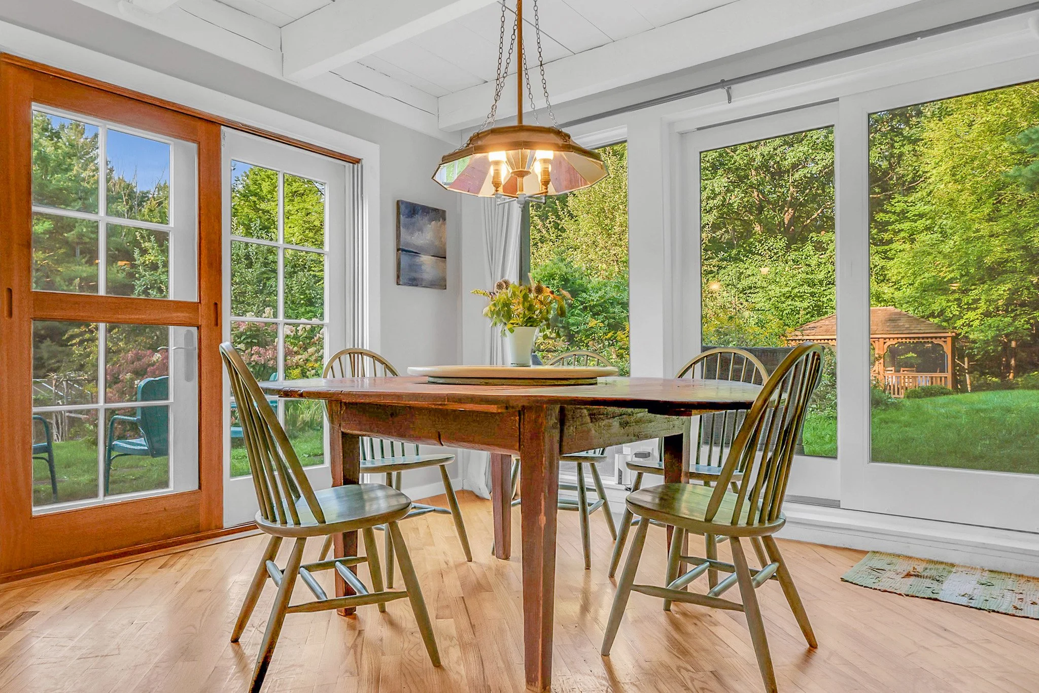 Dining room with wooden table, six matching chairs, large windows showing green trees outside, and a chandelier hanging above the table.