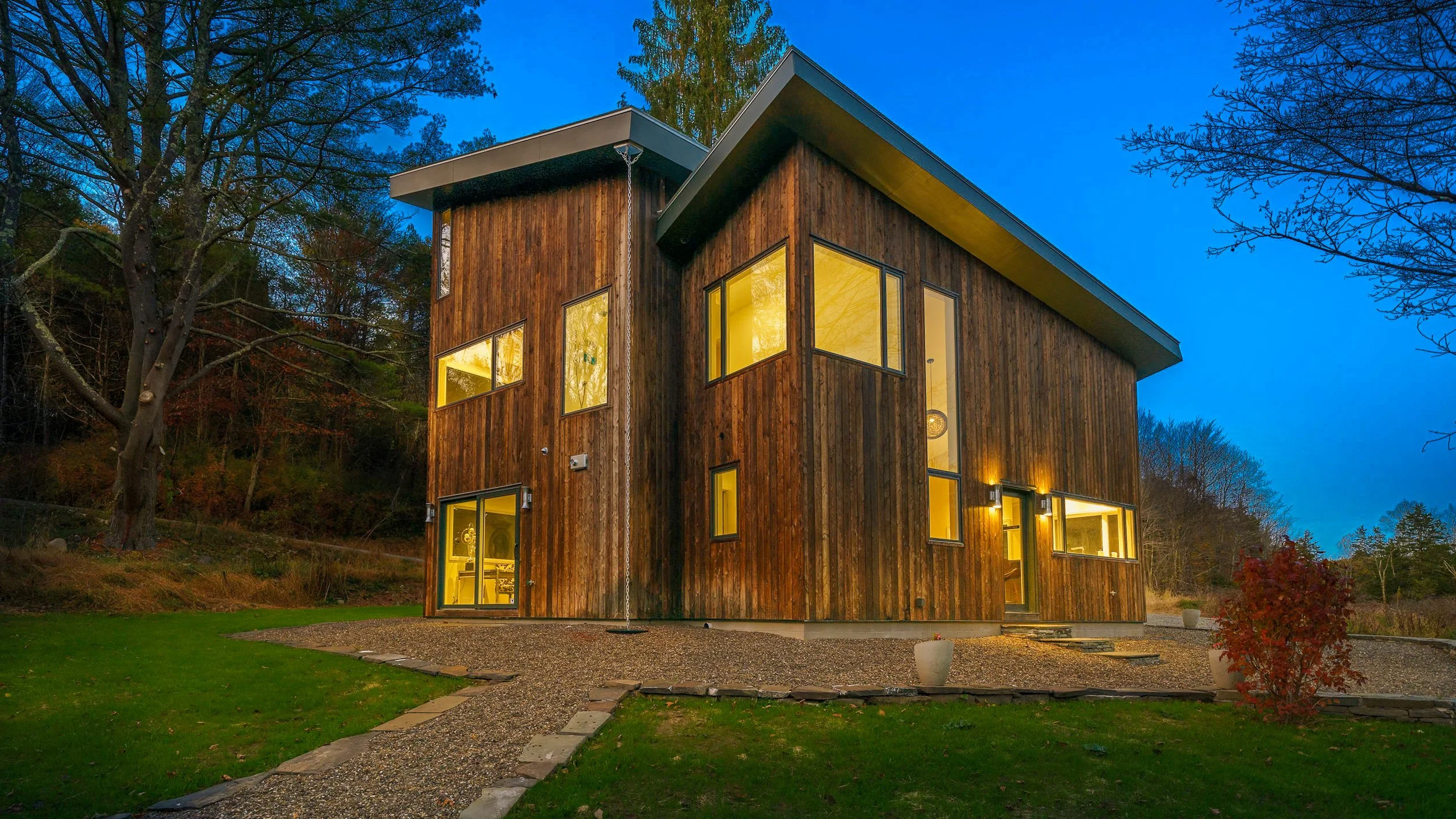 Modern wooden house at dusk with illuminated windows, surrounded by trees and a garden with gravel paths.