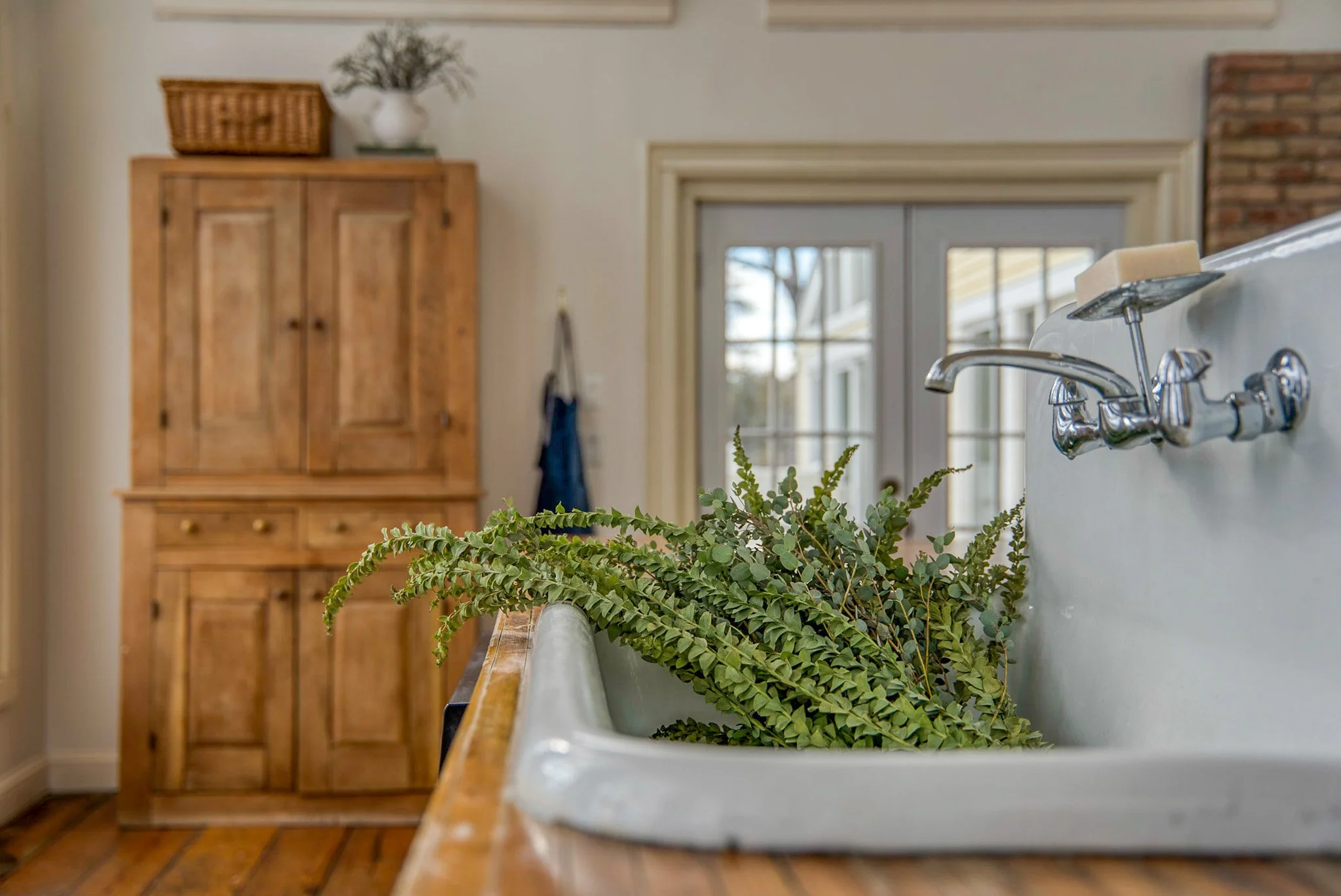 Kitchen sink with green leafy plant inside, wooden countertop, wooden cabinet, glass door in the background, and a brick wall to the right.
